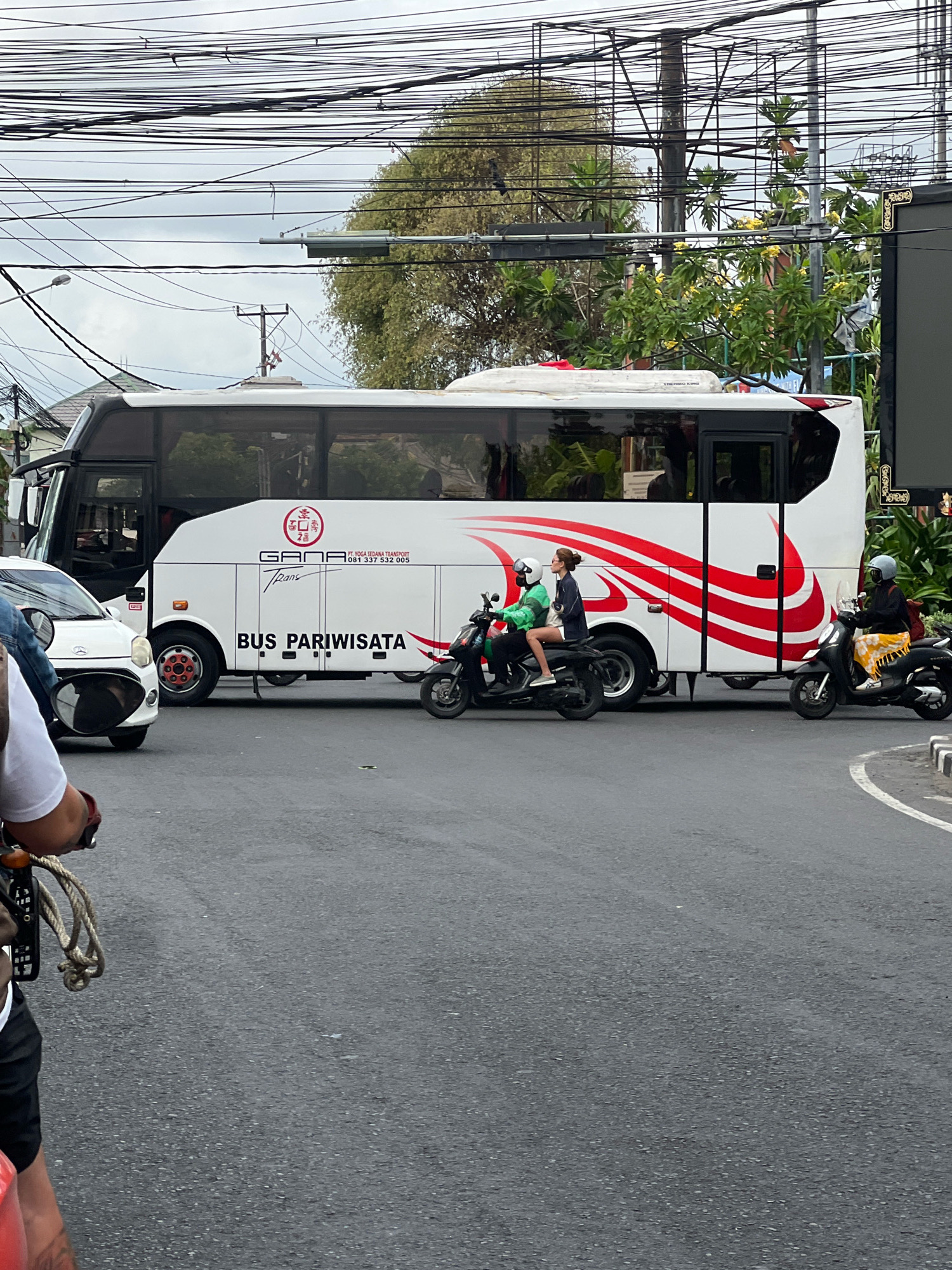 The image depicts a street scene in Denpasar, Indonesia, likely during the daytime under an overcast sky. A white tourist bus, labeled "BUS PARIWISATA," with red decorative stripes, is parked or moving slowly on the road. On the bus, a logo with red Chinese characters is visible. To the left of the bus, a white car is partially visible. Two motorbikes are also present. The first motorbike, a black scooter, carries a woman wearing a green jacket and shorts, and a man in front of her. The second motorbike, also a black scooter, is ridden by a woman with a yellow sarong. Power lines are a prominent feature of the scene, crisscrossing the sky. Trees with green foliage and some yellow flowers are visible in the background, along with a glimpse of a building with a dark facade. The road surface is asphalt, with a white line marking the edge on the right side.