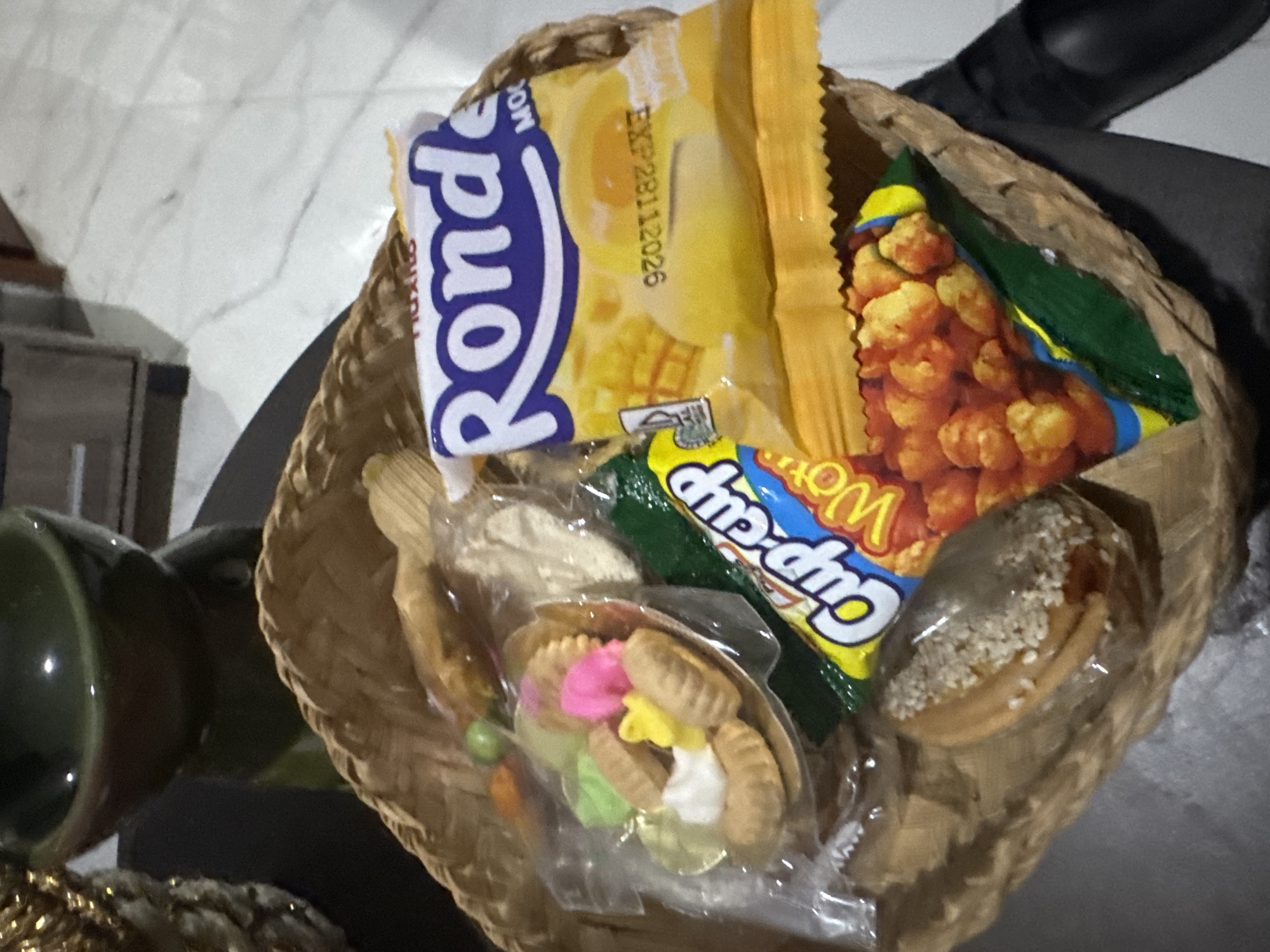 The image shows a close-up, top-down view of a woven basket filled with various snacks. The basket is situated on a dark, reflective surface, possibly a table, with a blurred background that includes a white tiled floor and furniture. The snacks include a bag of "Ronde" cookies, a package of "Cup-cup" candy with the word "Worm" visible, a small bowl containing colorful candies and cookies, and a single cookie topped with sesame seeds. The overall lighting suggests an indoor environment. No people are visible in the image. The image appears to have been taken in Sukawati, Indonesia, as indicated by the presence of a "Cup-cup" candy, a popular snack in Indonesia.