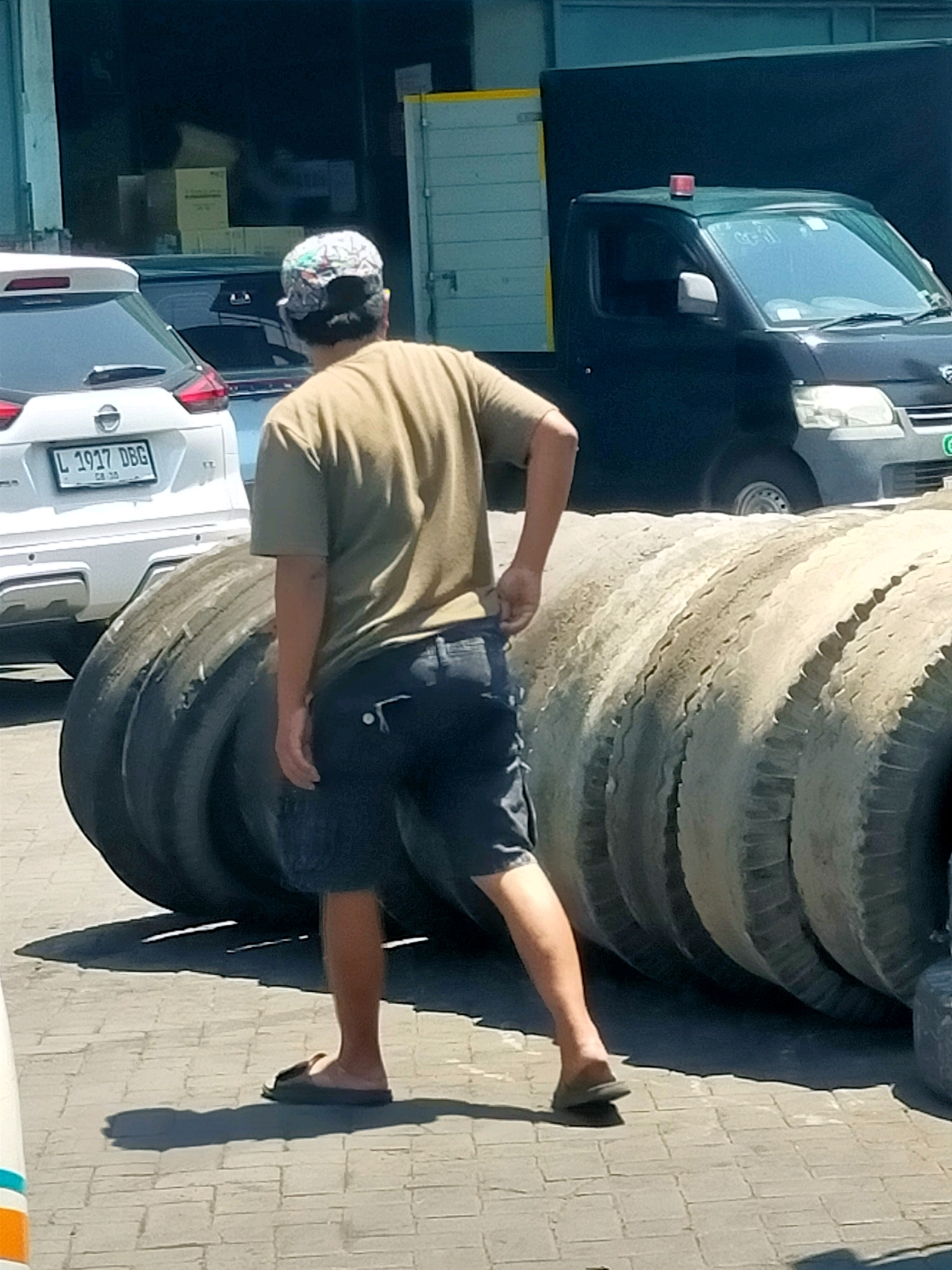 A man wearing a patterned baseball cap, a tan t-shirt, and denim shorts stands in front of a stack of large tires in what appears to be an industrial or commercial area. He is seen from behind, with his right hand near his shorts pocket and his left arm slightly bent. The tires are worn and dusty, suggesting they are old or have been used extensively. In the background, a white SUV with an Indonesian license plate (L 1937 DBG) is parked, and a dark-colored truck with a red emergency light on its roof is also visible. The ground is paved with interlocking bricks. The scene suggests a location possibly related to transportation, tire services, or a warehouse in Surabaya, Indonesia, given the context.