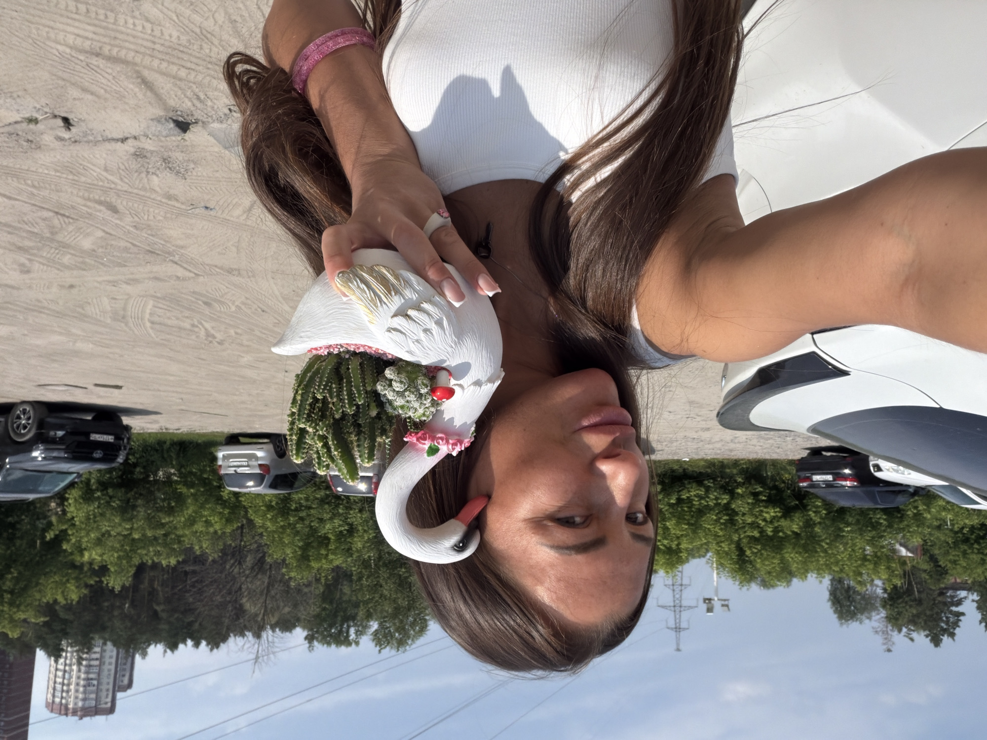 A woman is taking a selfie outdoors in what appears to be a parking lot or industrial area. She is holding a white swan-shaped planter containing succulents and a small red mushroom decoration. She is wearing a white tank top and has long brown hair. In the background, there are several cars parked, including a white car, a black sedan, a silver minivan, and a black SUV. Beyond the cars, there is a line of green trees and a few buildings, including a tall residential building. There are also power lines and a high-voltage tower visible in the distance. The ground is paved with dirt and sand, showing tire tracks. The sky is clear and blue. The overall impression is a snapshot of a sunny day in a semi-urban or suburban setting.