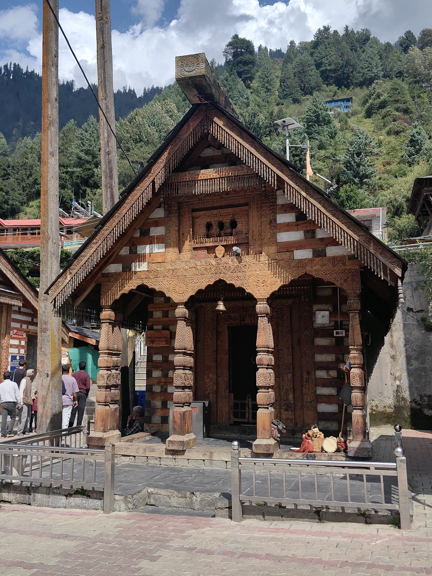 The image captures the exterior of the Vashisht Temple in Vashisht village, India. The temple features intricate wooden carvings on its facade, including decorative pillars and roof details. The architectural style is traditional Himachali. In the foreground, a metal railing separates the temple entrance from a paved walkway. Several people are visible near the entrance, some walking and others standing. One person is seated in the doorway. The temple is situated on a hillside covered with lush green trees, indicating a mountainous or hilly region. The sky is partly cloudy. Behind the temple, a blue building is visible further up the hill. There are also utility poles and wires present in the scene. The overall impression is one of a cultural and religious site set within a natural landscape.