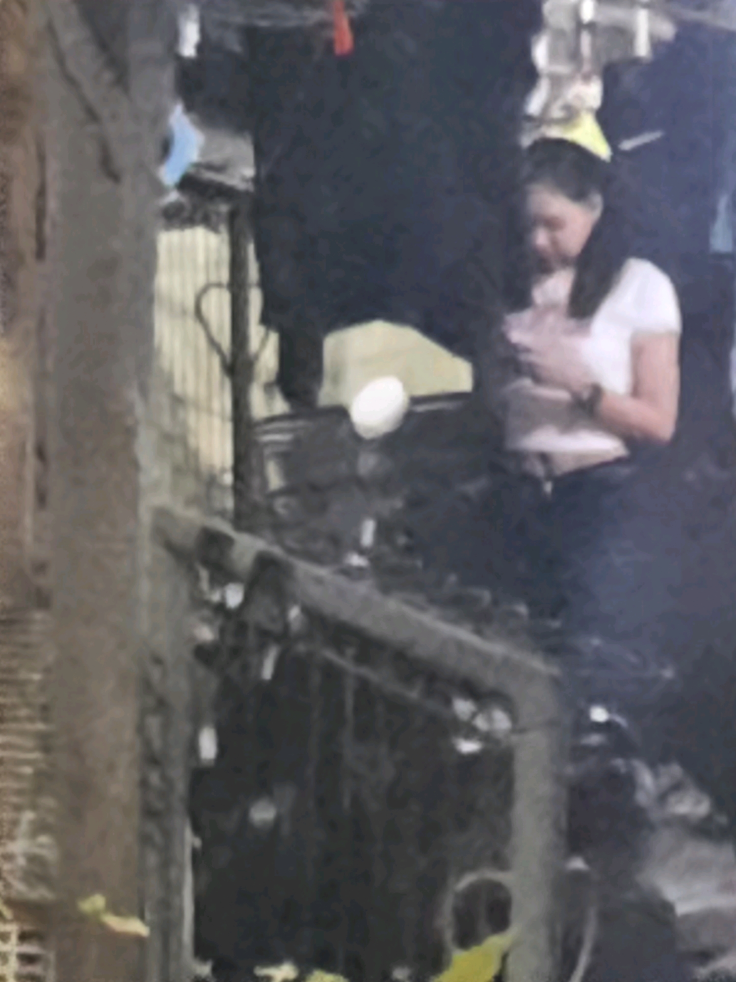 The image depicts a blurry outdoor scene in East Jakarta, Indonesia, with a woman in a white t-shirt and dark pants standing in the foreground. She is looking down at her hands, which are held in front of her chest. Behind her, a dark, possibly metallic structure is visible, with what appears to be a shopping cart or trolley. The background is indistinct, with hints of urban elements. The lighting suggests it might be evening or nighttime, with some artificial light sources present.