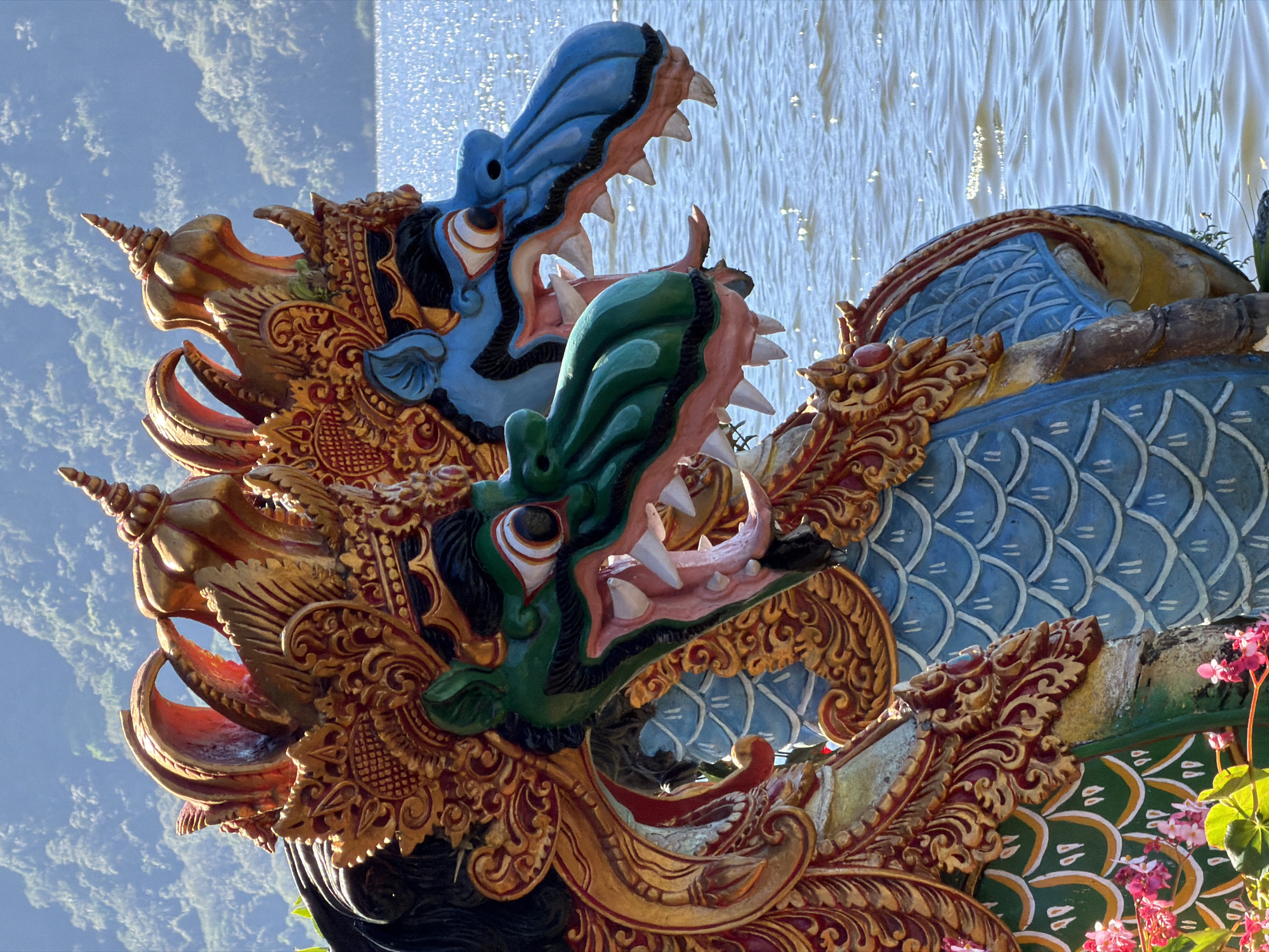 Two ornate dragon statues stand side-by-side on the edge of a body of water, likely a lake or reservoir. The statues are vibrantly colored, with the front dragon featuring a green head and intricate golden and red detailing around its face and headdress. Its mouth is open, revealing sharp white teeth and a pink tongue. The dragon behind it has a blue head and similar ornate decorations. The statues are positioned in front of a sunlit body of water, with ripples reflecting the light. In the background, a hazy, green, tree-covered mountainside rises under a clear, blue sky. The scene is illuminated by bright sunlight, casting strong shadows and highlighting the textured surfaces of the statues. The presence of flowers and foliage in the foreground suggests a natural or landscaped setting. The dragon statues are reminiscent of those found at the Ulun Danu Beratan Temple, a significant landmark located in the village of Candikuning, Baturiti, Bali, Indonesia, known for its water temple architecture and scenic mountain backdrop.