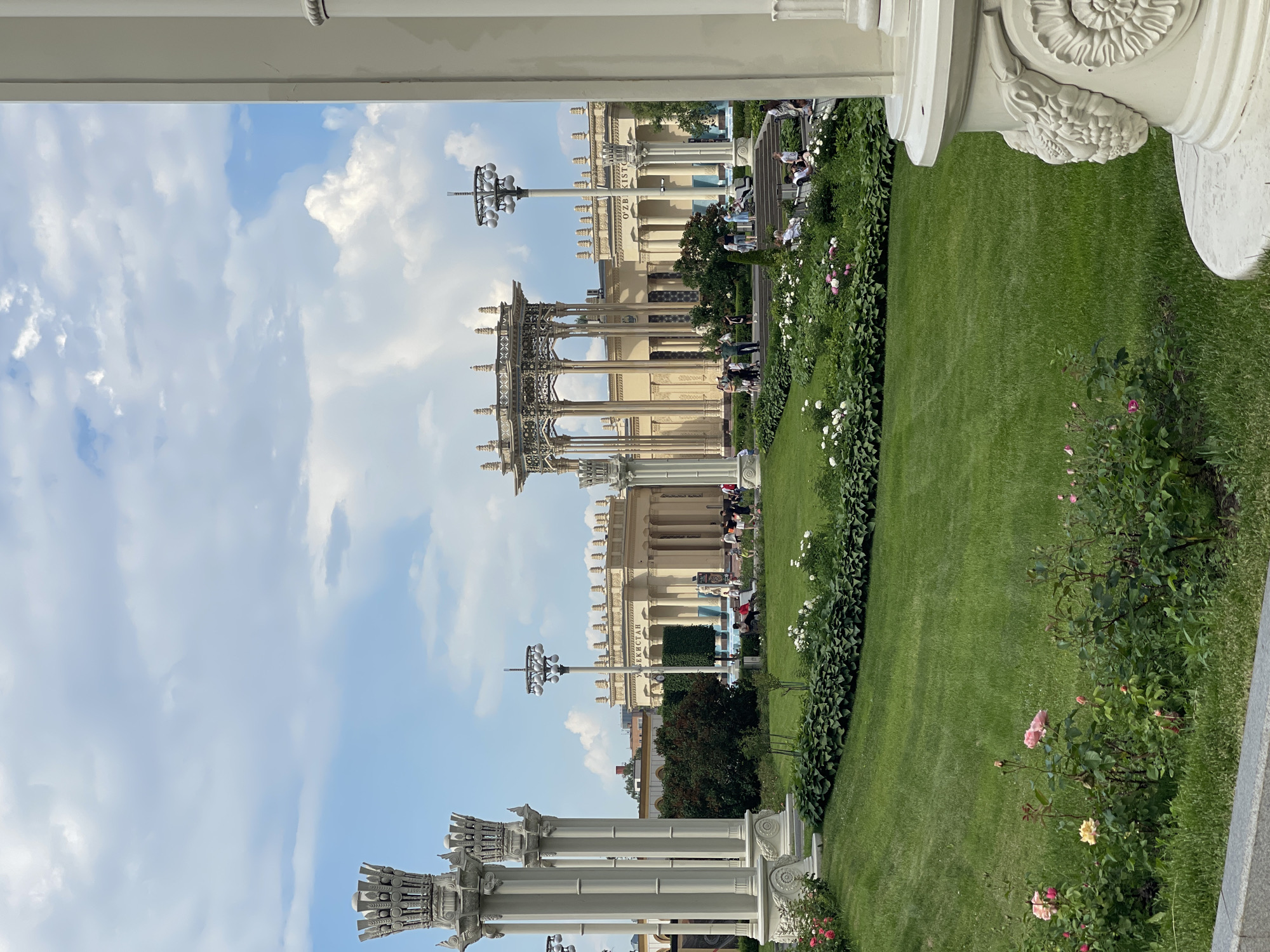 A medium shot captures a sunlit park with lush green grass and flowering bushes in the foreground. In the midground, a grand pavilion with intricate metalwork and classical architecture stands out, flanked by a long, colonnaded building with arched windows. The building bears Cyrillic lettering on its facade, indicating "UZBEKISTAN". Several people are scattered throughout the park, some sitting on benches or walking along pathways. The sky above is a vibrant blue, adorned with fluffy white clouds, suggesting a pleasant day. The overall scene conveys a sense of grandeur and public gathering in an outdoor setting.