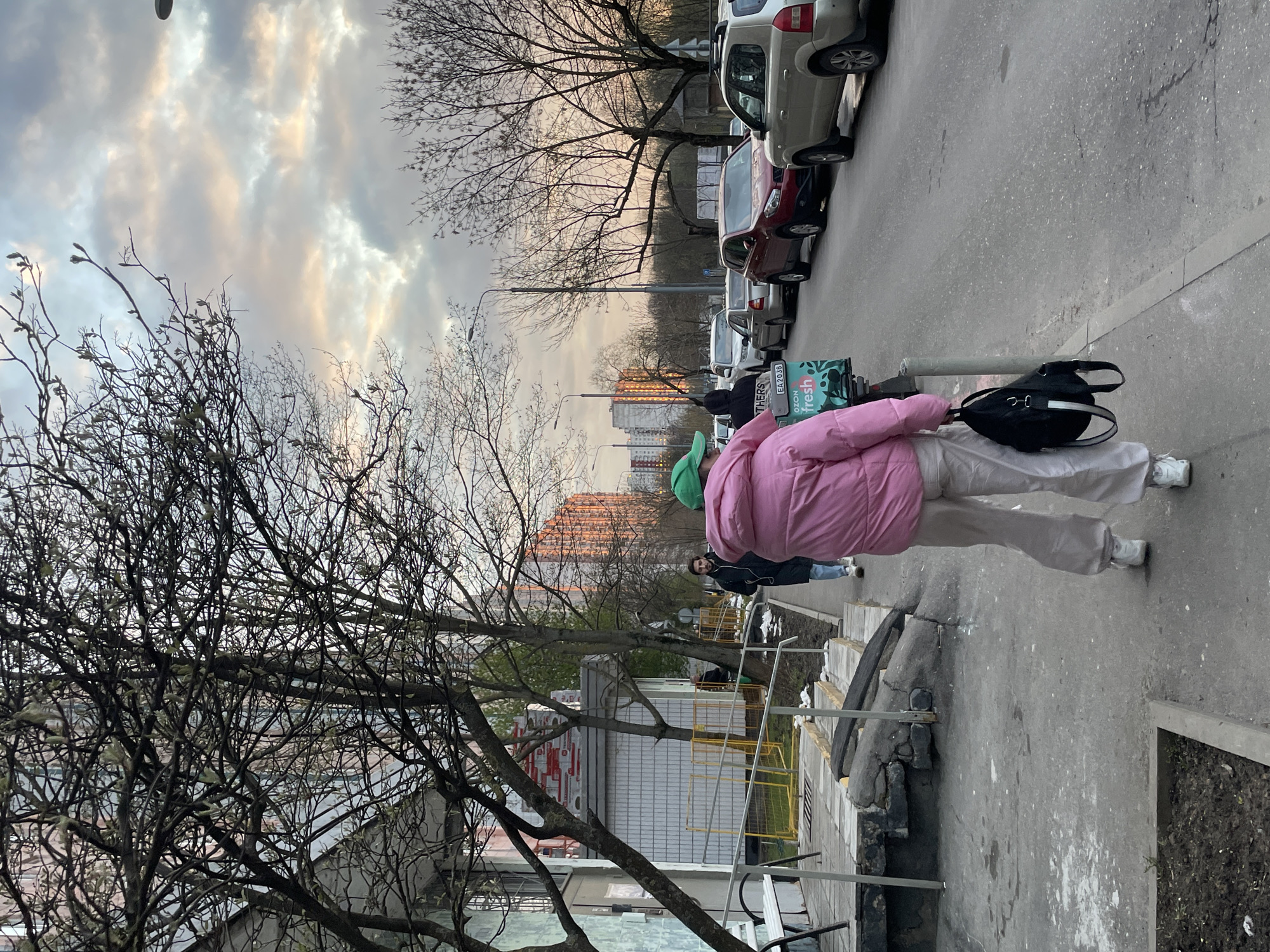 The image captures an urban street scene in Moscow, Russia, likely during late afternoon or early evening due to the warm, golden light reflecting on the clouds and distant buildings. The sky is partially overcast with dramatic, streaky clouds displaying hues of grey, white, and soft orange.
In the foreground, a person is seen from behind, wearing a prominent pink puffer jacket, light-colored wide-leg trousers, white sneakers, and a green cap. They are standing or walking next to a green and black delivery box or scooter, on which a black backpack with white accents is placed. The delivery box clearly displays the text "ozon fresh" in white lettering on a green background, along with other visible alphanumeric characters such as "EA 2036" and fragments like "THERS" on what appears to be a license plate or identification sticker.
The street extends into the mid-ground, lined with several parked cars on the right, including a light-colored SUV, a dark red sedan, and other vehicles. Deciduous trees, mostly bare with some early budding leaves indicating early spring, frame the left side of the street and stand between the road and the buildings. A second person, dressed in a dark jacket and blue jeans, is visible further down the sidewalk on the right, facing slightly towards the camera.
In the background, tall residential apartment buildings rise, with some facades illuminated by the setting sun. The ground consists of paved sidewalks and an asphalt road, showing some cracks and wear. Yellow guard rails are visible near a structure in the mid-ground, possibly indicating a construction or renovation site. The overall scene depicts everyday urban life under a dynamic sky.