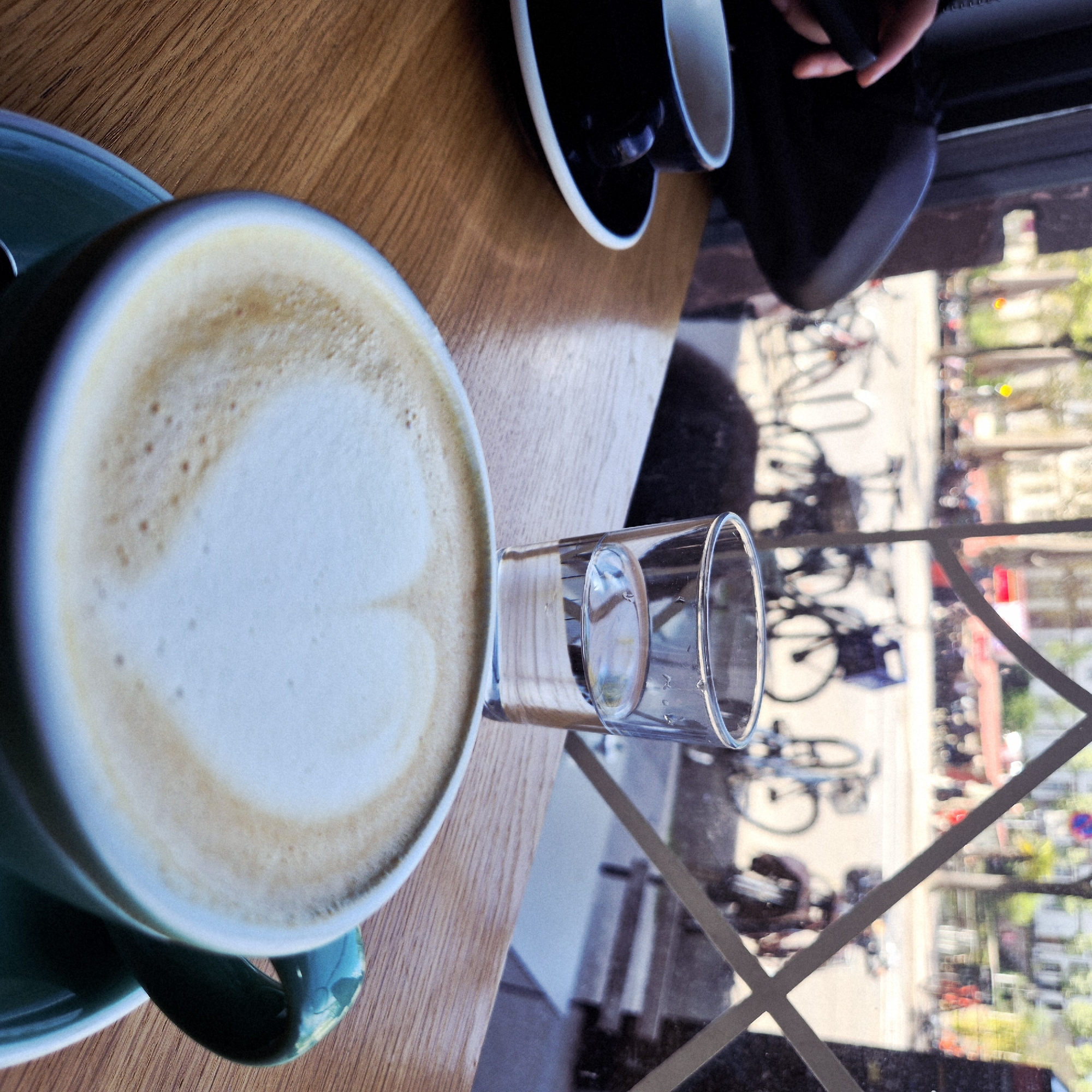 The scene depicts a wooden table with a turquoise cup of coffee and a glass of water next to it. Another cup of black coffee and a purse are also on the table. A street scene is visible through a window, revealing parked bicycles, trees, and people in Strasbourg, France.