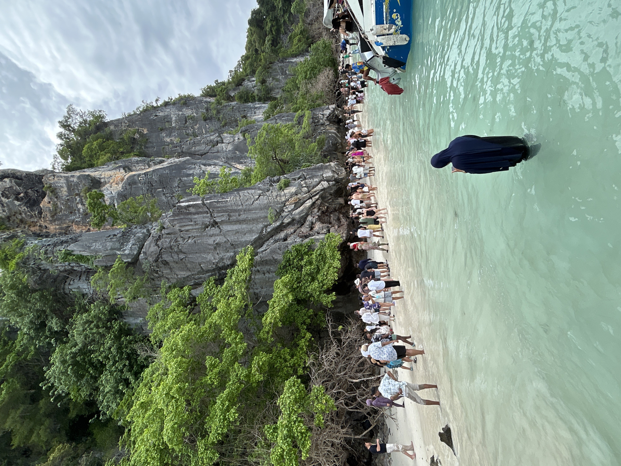 A group of tourists stands on a sandy beach in shallow, clear turquoise water. They are gathered near the base of a large, gray limestone cliff covered in lush green trees and vegetation. A few people are wading in the water, while others stand on the sand. One person, wearing a dark blue hijab and covering, stands in the water looking at their phone. A blue and white boat is docked to the right of the image, with a few people on board. The sky is cloudy and overcast. The scene appears to be a tourist destination, likely on an island near Ko Phi Phi Leh, Krabi province, Thailand, known for its dramatic limestone cliffs and clear waters.