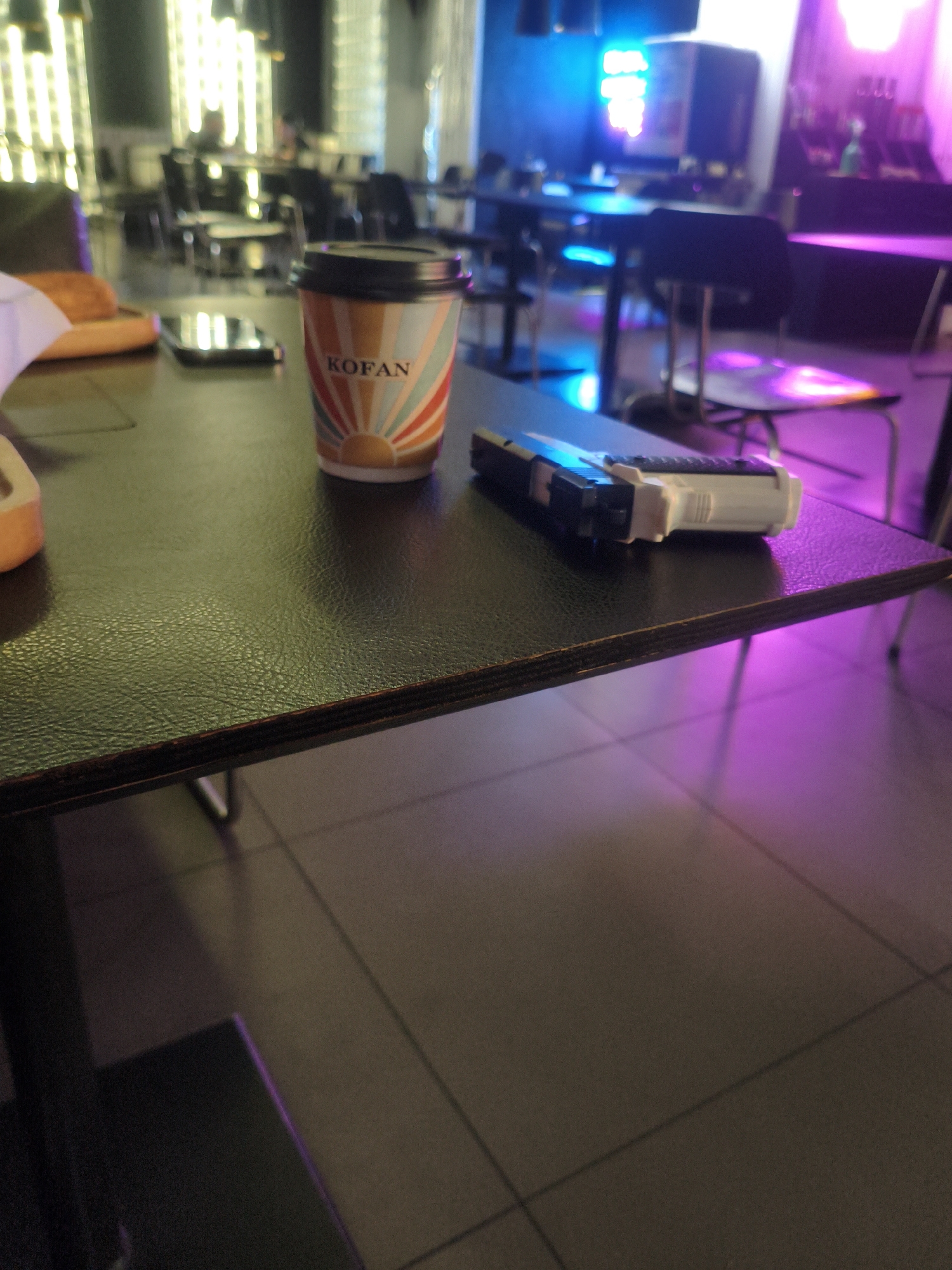 The image depicts a dark-colored table in what appears to be a dimly lit establishment, possibly a cafe or bar, with a blurred background of patrons and neon lighting. On the table, there is a disposable coffee cup with a sunrise design and the word "KOFAN" printed on it. Adjacent to the cup is a toy gun, appearing to be a replica of a handgun. The lighting in the background suggests a vibrant atmosphere with blue and pink hues. The floor is tiled. The overall scene appears to be a still-life arrangement on the table, with no discernible actions or interactions occurring. The location is identified as Kremenchuk, Ukraine.