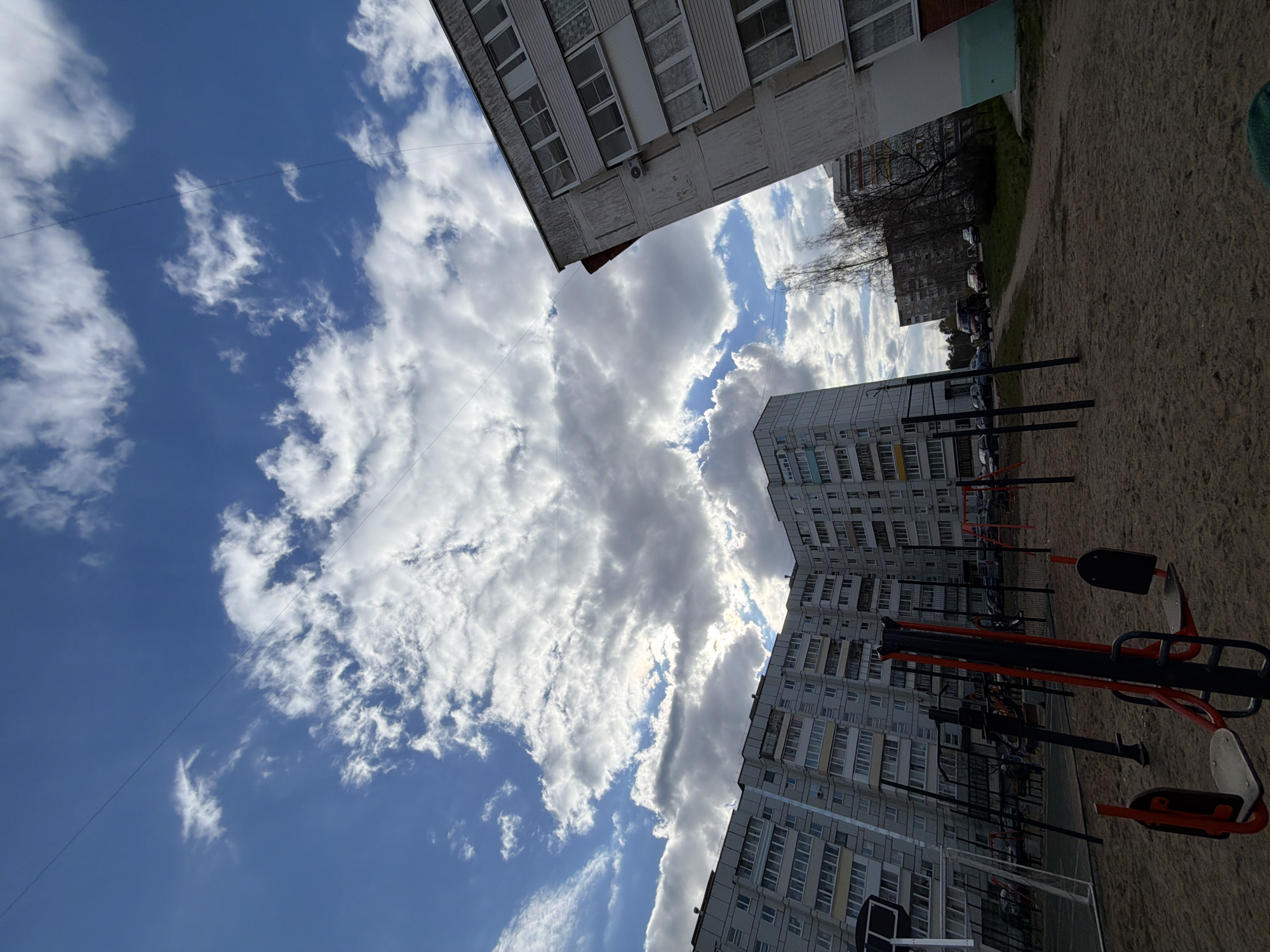 The image depicts an outdoor residential scene, likely a courtyard or common area between apartment buildings in Chaikovsky, Russia. The viewpoint looks across a ground area and upwards towards the sky and surrounding structures.
Several large, multi-story apartment buildings are prominent. One building's upper portion is visible on the left side of the frame, featuring multiple windows and a somewhat weathered facade. A much larger apartment complex dominates the right side and stretches into the background, characterized by numerous rows of windows and a gray, utilitarian architectural style typical of the region.
In the foreground and midground, a sandy or dirt-covered area contains red and black outdoor exercise equipment, along with part of a basketball hoop and net. In the distance, behind the buildings, bare trees and several parked cars are visible, suggesting a season other than summer, such as late autumn or early spring.
The sky is a significant element, showcasing patches of clear blue amidst an expanse of large, bright white and darker grey cumulus and stratocumulus clouds. Sunlight is clearly breaking through the clouds, indicating a bright daytime setting. Thin wires or power lines traverse the sky.
No people are present in the image, and no specific activity or event is taking place, rendering a tranquil and ordinary urban landscape. No discernible text is visible anywhere in the scene. The architectural style provides a strong location cue consistent with the provided context of Chaikovsky, Russia.