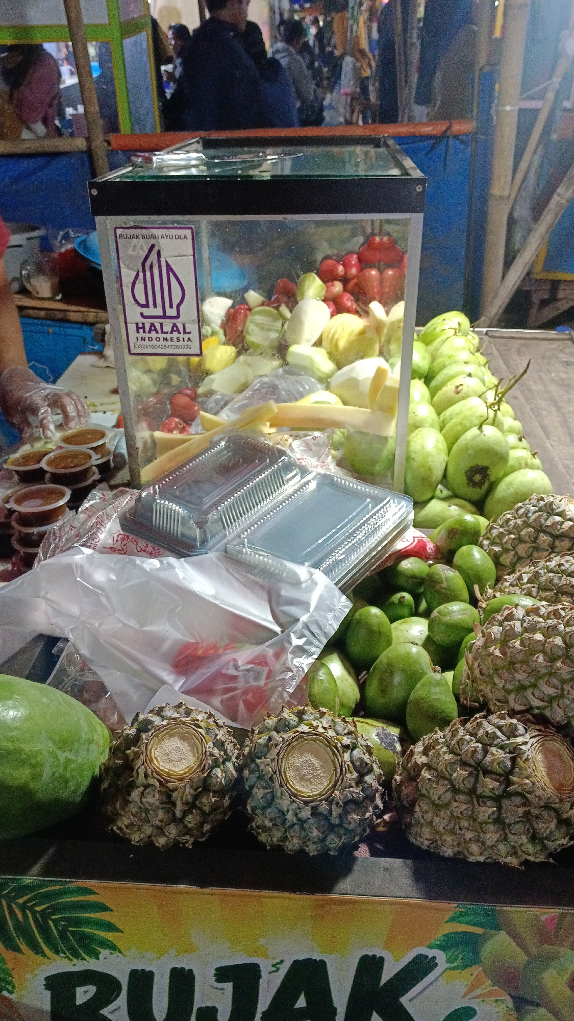 The image shows a food stall selling fruit salads, likely in Tajurhalang, Indonesia. In the foreground, there are three pineapples, one papaya, and several green fruits that appear to be avocados or similar. Behind these, there are transparent containers filled with chopped fruits like mangoes, tomatoes, and possibly guava. A glass display case contains more prepared fruit, along with a purple sticker indicating "HALAL INDONESIA." Several small containers of sauce are stacked to the left, and a person wearing a transparent glove is reaching towards them. In the background, other stalls and people are visible, suggesting a busy marketplace environment. The overall scene depicts a street food vendor preparing and selling fruit-based dishes.