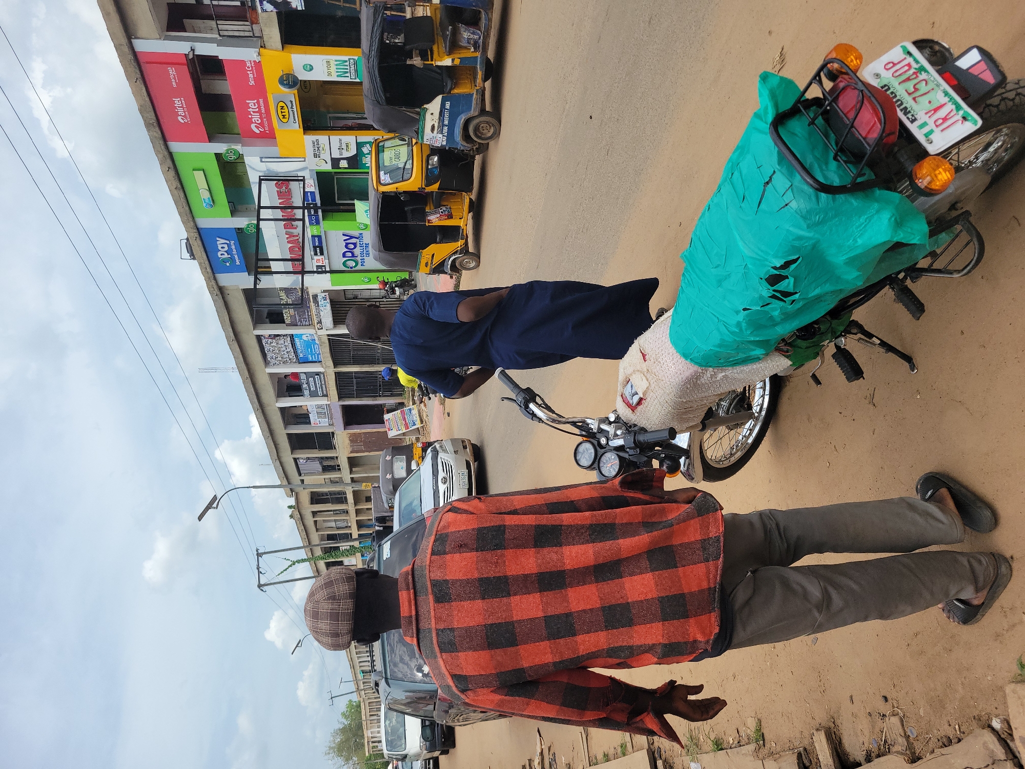 The image depicts a street scene in Egbagu, Nigeria. Two men stand next to a parked motorcycle with a green plastic cover on the back. One man wears a red and black checkered shirt and a cap. The other man wears a blue shirt. The motorcycle has a license plate that reads "JRV-7540P" and indicates "ENUGU COAL CITY STATE." In the background, there are buildings with signage for various businesses, including Airtel, Glo, MTN, and Opay. Several "Keke Napep" auto rickshaws are parked along the road. A street light and power lines are also visible. Several cars are passing.