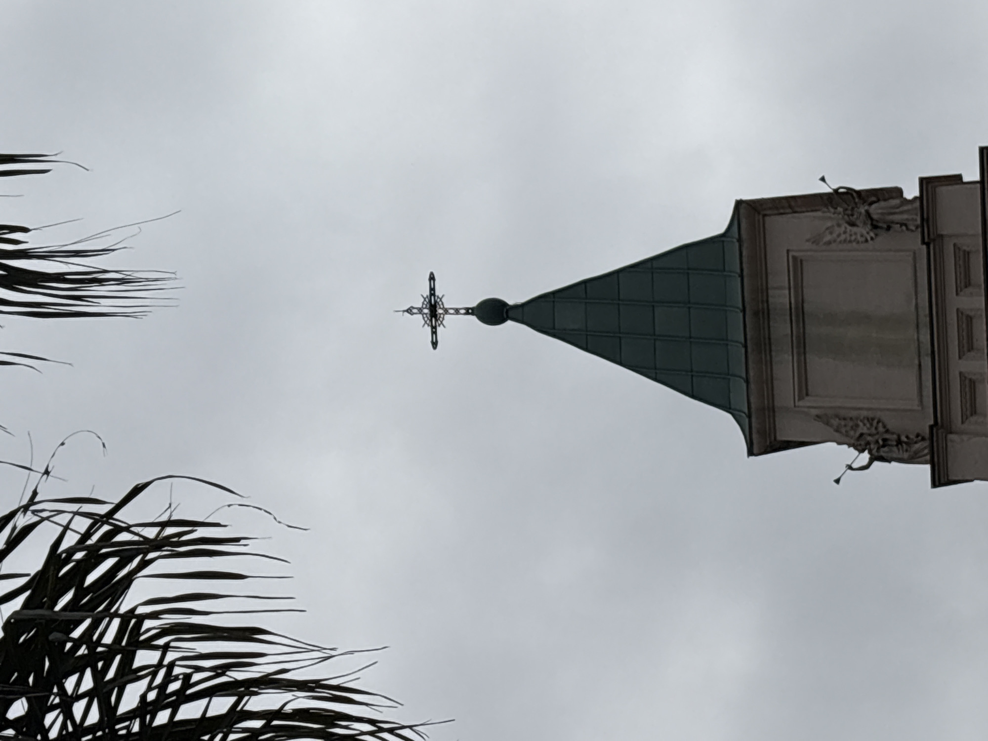 The image shows the upper portion of a church tower against a cloudy sky. A prominent feature is the church's steeple, which is covered in dark green, tiled material, culminating in a metallic cross at its apex. The cross is ornate, with intricate detailing. Below the steeple, the tower has a light-colored facade with decorative architectural elements, including what appear to be statues of angels on either side, each holding a horn. Palm fronds are visible in the upper left corner, framing the scene. The overall impression is one of a religious building on a somewhat overcast day. This is the spire of the Igreja da Nossa Senhora do Brasil, located in the Morumbi neighborhood of São Paulo, Brazil.