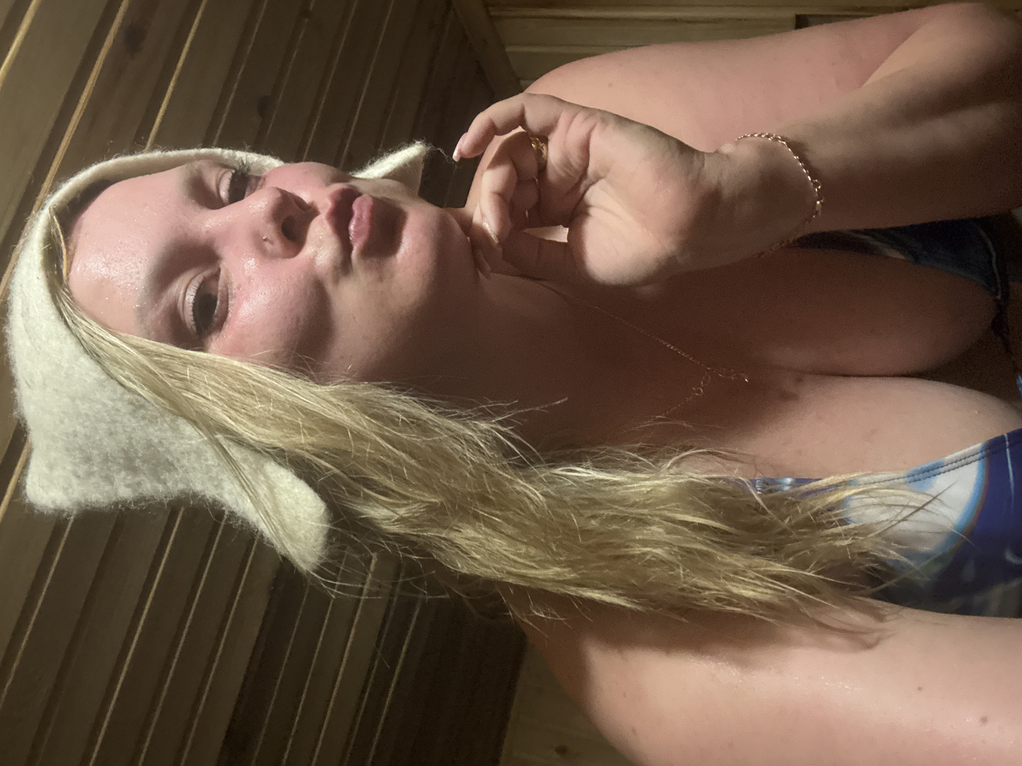 A woman with blonde hair and a white wool hat is reclining in a sauna with wooden walls. She has a fair complexion, and is wearing a blue and white top. She is touching her neck with her right hand, which has a gold ring on one of her fingers. A gold chain and a bracelet adorn her. The location is Dubovaya, Russia.