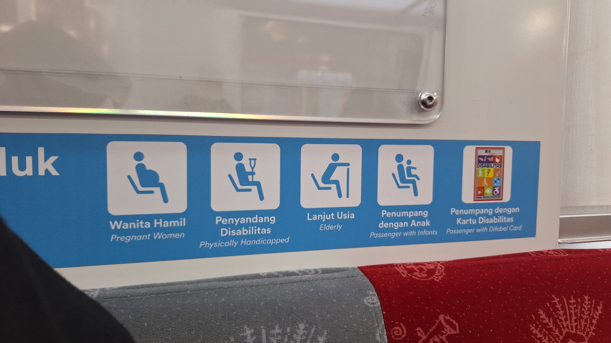 The image shows a close-up view of a blue sign mounted on the interior wall of a public transport vehicle, likely a train or bus, in Cikarang, Indonesia. The sign features several icons with accompanying text in Indonesian and English, indicating priority seating for specific passenger groups. From left to right, the icons and text are:
1. A pregnant woman sitting. Text: "Wanita Hamil" / "Pregnant Women".
2. A person sitting with crutches. Text: "Penyandang Disabilitas" / "Physically Handicapped".
3. An elderly person sitting. Text: "Lanjut Usia" / "Elderly".
4. A person sitting with a child on their lap. Text: "Penumpeng dengan Anak" / "Passenger with Infants".
5. A small card with various icons and text that reads "DISABILITAS" and "Passenger with Difabel Card".
The sign is positioned above a row of seats, part of which is visible at the bottom of the image, showing red and gray upholstery. The lighting suggests an indoor environment, possibly during daylight hours. The overall presentation is clean and informational, typical of public transportation.