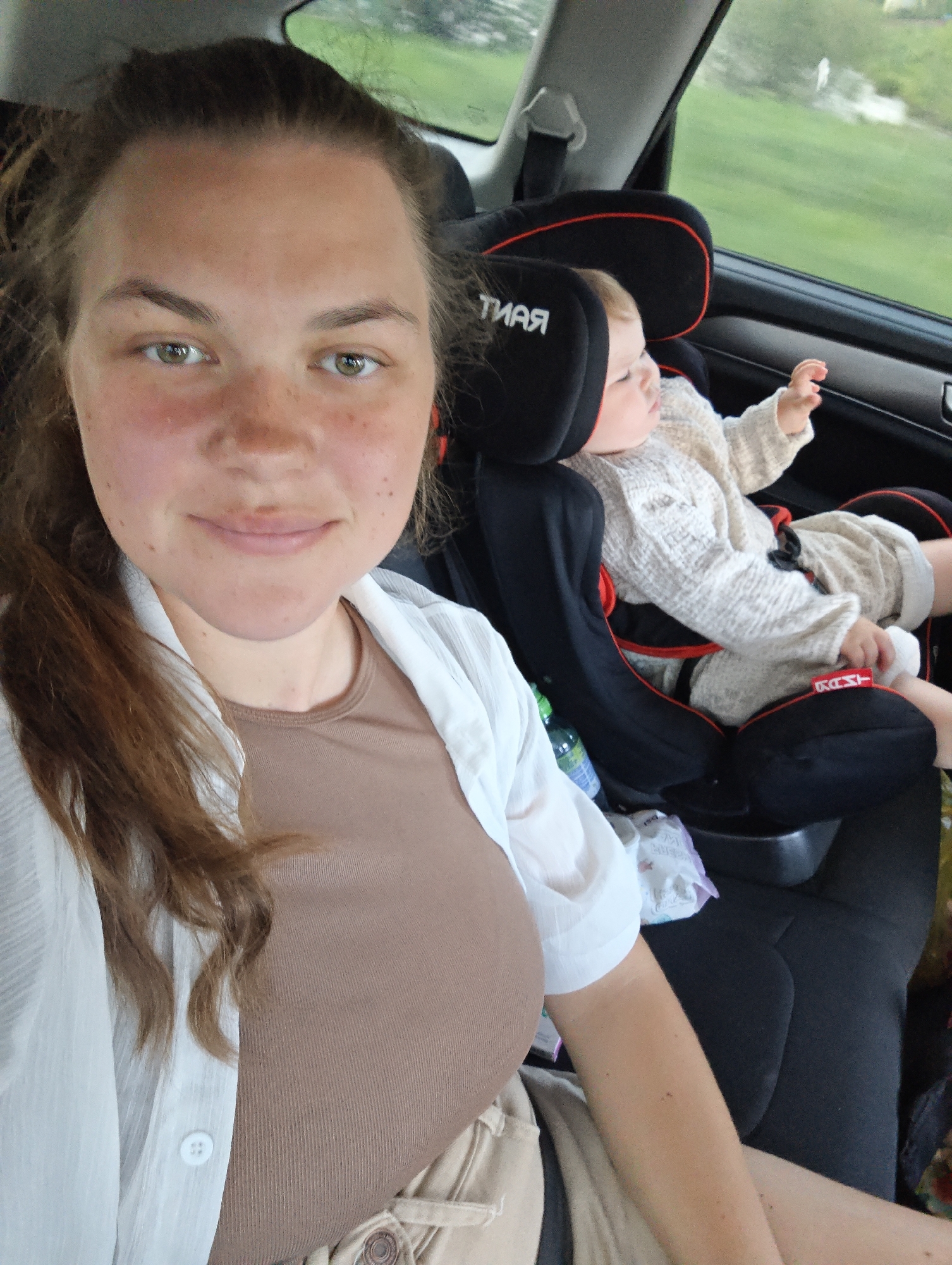 A woman is taking a selfie in the back seat of a car. She has long brown hair and is wearing a brown shirt and a white button-down shirt. A baby is secured in a car seat beside her, looking out the window. A bottle of water and a package of wipes are visible near the car seat. The window shows greenery outside. The location Фоки, Russia, is not discernible from the image.