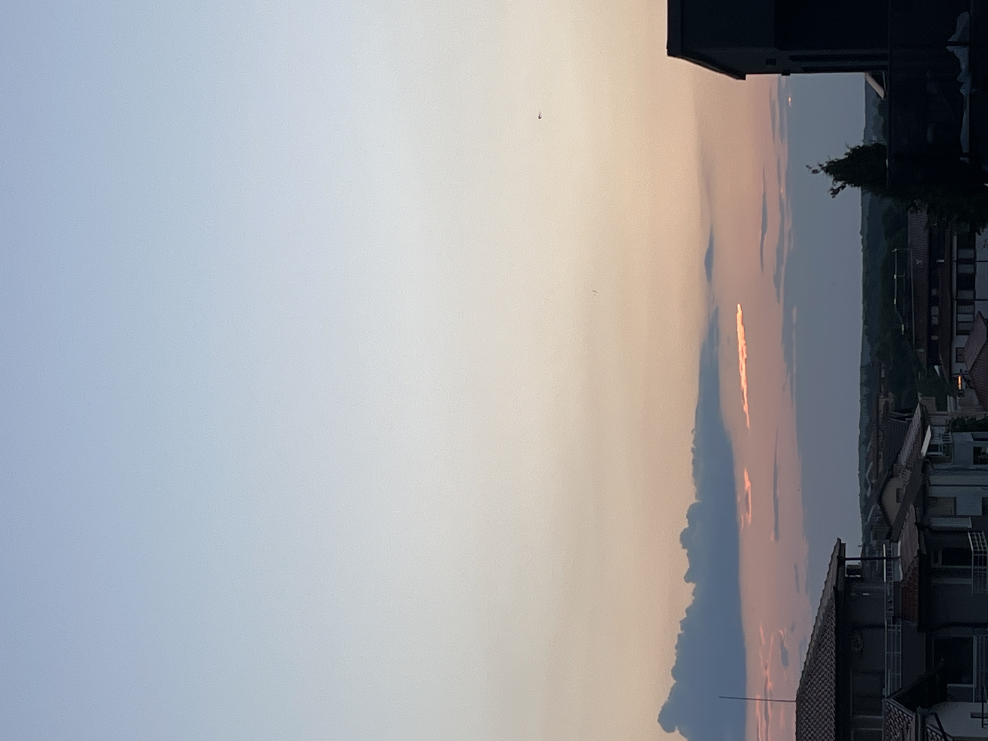 The image depicts a panoramic view of the city of Lozenets, Bulgaria, during what appears to be sunset or dusk. The sky transitions from a pale blue at the top to soft hues of pink and orange near the horizon, indicating the presence of clouds illuminated by the setting sun. A few dark clouds are visible in the middle ground, with some streaks of orange light piercing through them. In the foreground, the rooftops of several buildings are visible, characterized by terracotta tiles and modern architectural designs. A tall, dark, rectangular building is positioned on the right side of the frame, partially obscuring the view of the distant landscape. A lone tree with dark foliage stands to the left of this building. The overall scene conveys a sense of tranquility and the beauty of a natural twilight phenomenon in an urban setting. There are no people or discernible actions captured in the image.