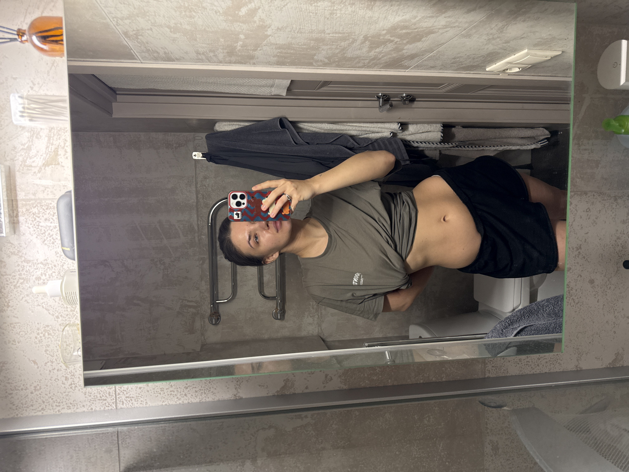 A young woman stands in a bathroom, taking a mirror selfie. She has dark hair pulled back and a neutral expression. She is wearing a khaki or olive green short-sleeved t-shirt, which is partially lifted to reveal her midriff and navel, and black shorts. She holds a smartphone with a distinctive colorful, patterned case featuring abstract shapes, possibly cartoon-like figures, in front of her face to capture the image. Text on her t-shirt appears to be "TYBO" or "TYBOO", visible as a mirror image.
The bathroom features textured light grey and beige tiles on the walls. In the reflection, a chrome heated towel rail with a dark grey towel and a folded white and blue striped towel are visible. Above the towel rail, there's a white ventilation grille and a white electrical outlet. To the left in the reflection, an orange glass diffuser with reeds and a white holder with cotton buds are placed on a surface. To the right, a white wall-mounted soap dispenser or similar item with a green accent is seen. The top edge of a white toilet is also visible in the lower right of the reflection. The overall lighting suggests an indoor setting, likely artificial. The mirror itself shows some smudges.