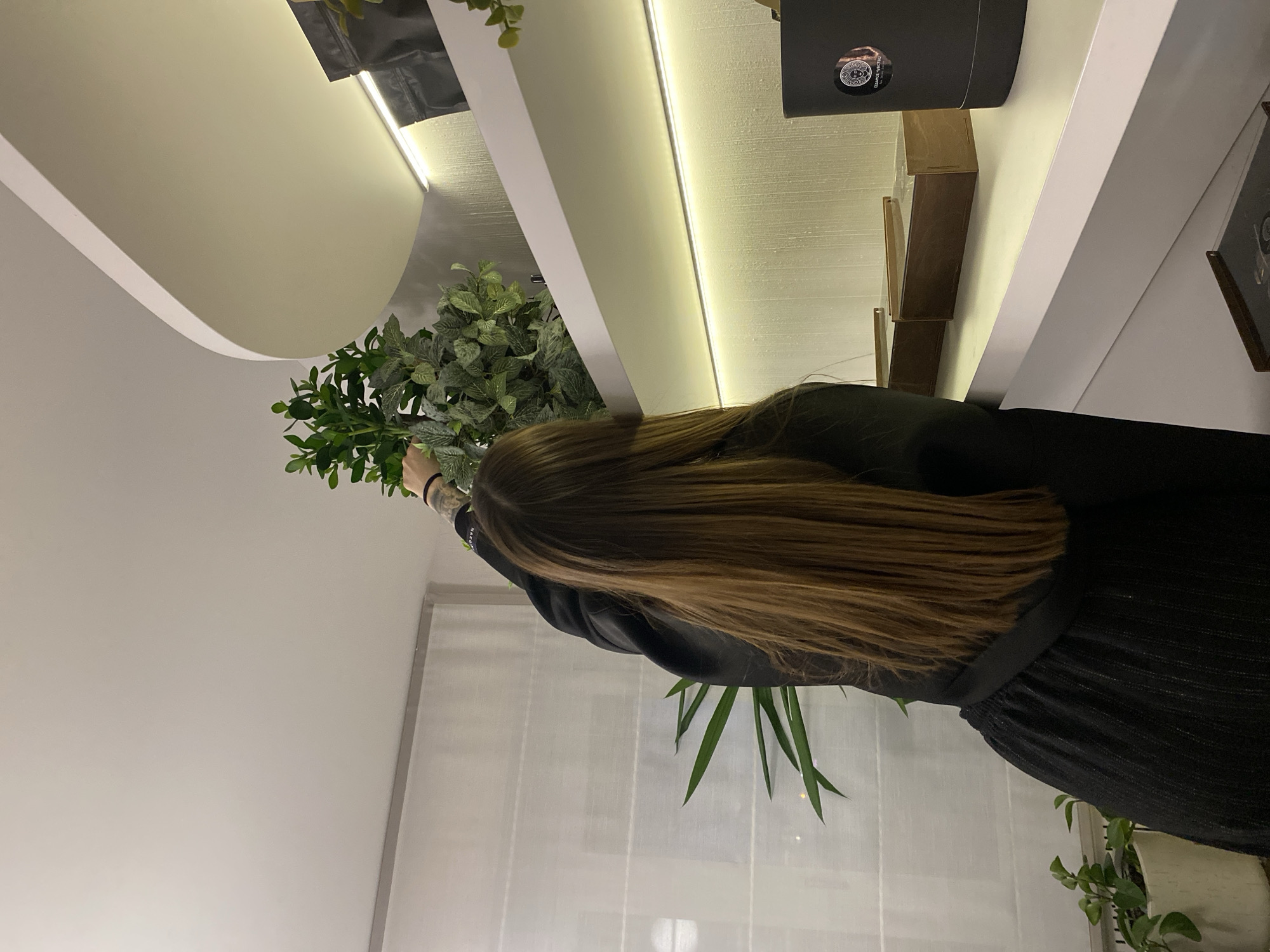 A person with long brown hair, wearing a black top and skirt, is tending to plants in a room. The plants are located near a window. The room has white walls and a white ceiling with recessed lighting. A shelf with a black box and other items is visible on the right side of the image. The location is indoors and the city of Votkinsk, Russia is not discernible from the image.