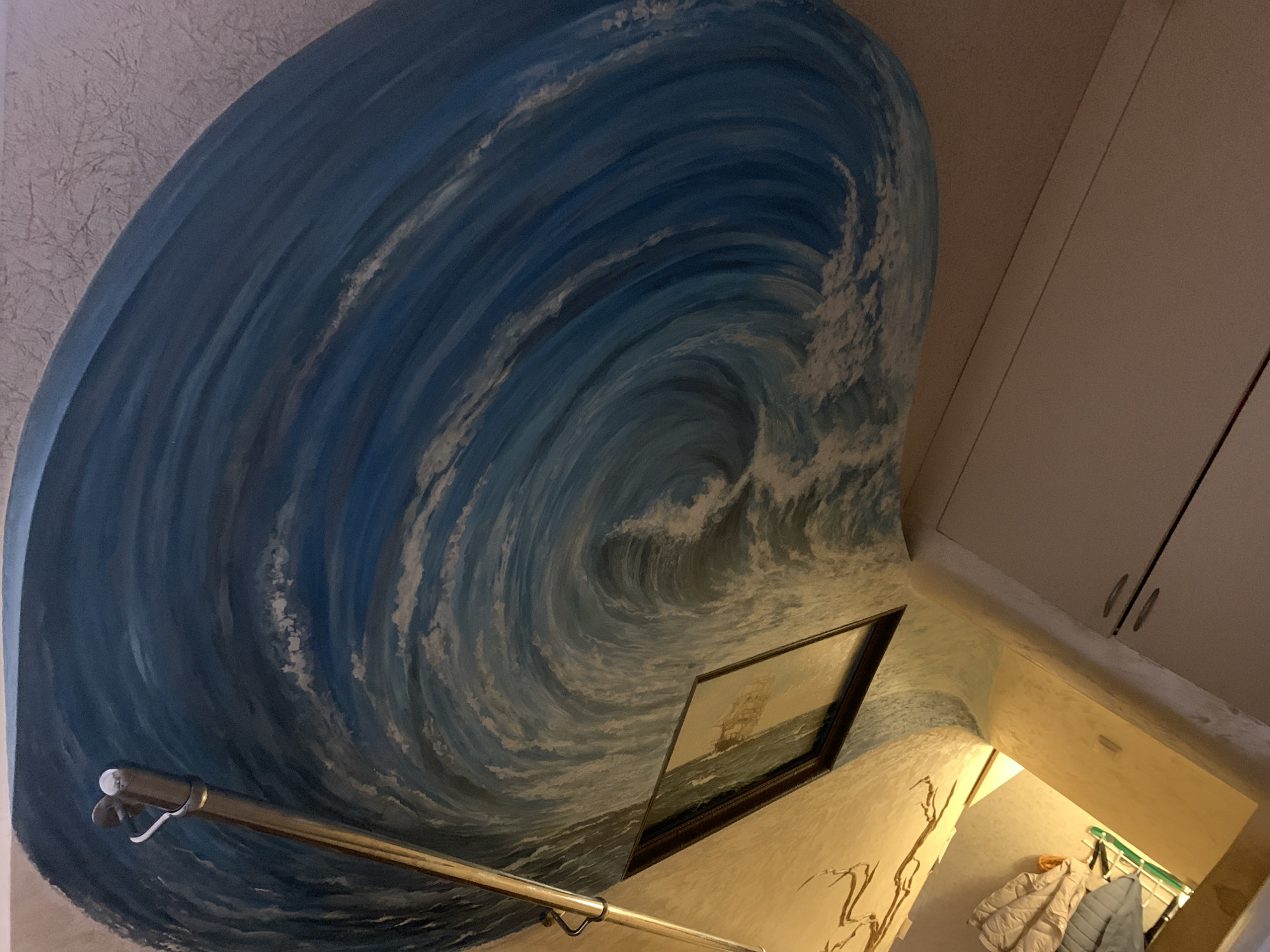 The image shows a corner of a room with a large mural of a swirling ocean wave painted on the wall. The mural dominates the left side of the frame, with shades of blue and white depicting the water's movement. Below the wave, a framed painting of a sailing ship is visible. A metal railing runs diagonally across the lower left of the image. The right side of the image shows a wall with built-in cabinets and a doorway leading to a brightly lit space. Clothes are hanging near the doorway. The image is taken at an upward angle. Severodvinsk, Russia, is not specifically identifiable from the image's content.