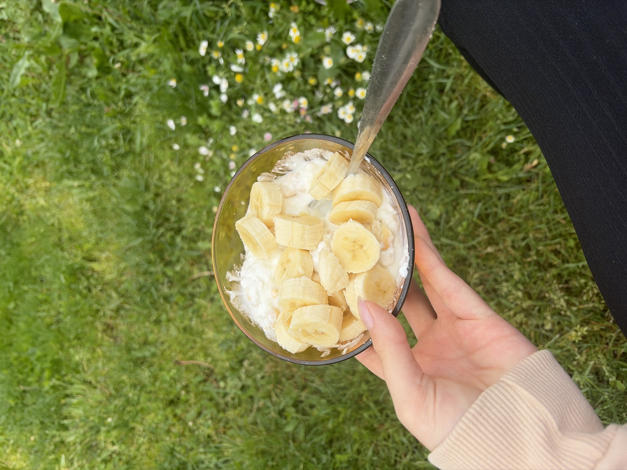 A high-angle shot depicts a hand holding a glass bowl filled with what appears to be cottage cheese and sliced bananas. A spoon is partially inserted into the mixture. The backdrop consists of a green lawn with scattered small white flowers. The lower portion of a person's leg clad in black fabric is visible on the upper right of the frame. The setting appears to be outdoors. There is no information to suggest that the photo was taken in the city of Brusen, Bulgaria.
