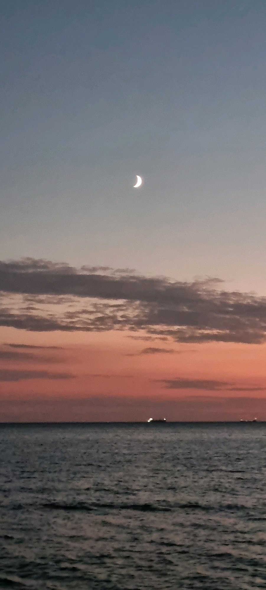 The image depicts a serene seascape at dusk. The sky transitions from a pale blue at the top to a warm gradient of pink and orange near the horizon, indicative of sunset or sunrise. A crescent moon is visible in the upper portion of the sky. A band of clouds stretches across the middle ground, with some appearing darker and more textured than others. Below the sky, the vast expanse of the sea is shown with gentle ripples on its surface. In the distance, near the horizon, several ships or boats are visible, their lights twinkling in the dimming light. One prominent ship with bright lights is positioned centrally, and another fainter one is to its right. The overall atmosphere is calm and tranquil.
This scene could be captured along the coastline of Bashkia Durrës, Albania, a region known for its Adriatic Sea beaches and popular seaside resorts. The presence of ships in the distance suggests proximity to shipping lanes or a port area, which is consistent with the geography of Durrës. The natural beauty of the sky and sea, combined with the distant signs of human activity, creates a picturesque and peaceful ambiance.