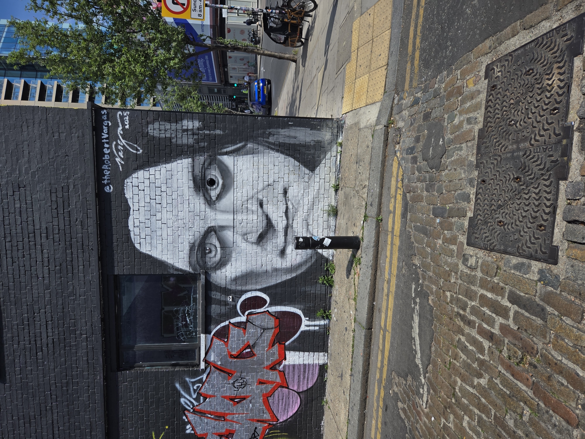 Here is a description of the image:
The image shows a wall mural of a person's face on a brick wall in an urban environment. Above the face is the signature of the artist, Robert Vargas, and the date 2015. A small window is embedded into the brick wall beneath the face. Below the face are some graffiti shapes in the colors red, grey, and purple. To the right of the wall is a brick sidewalk with a metal manhole cover embedded in it. A street with double yellow lines runs alongside the sidewalk. Across the street is a building with blue windows, as well as a small tree. In the background, there are road signs and bicycles. The image appears to have been taken in the city of London, United Kingdom.