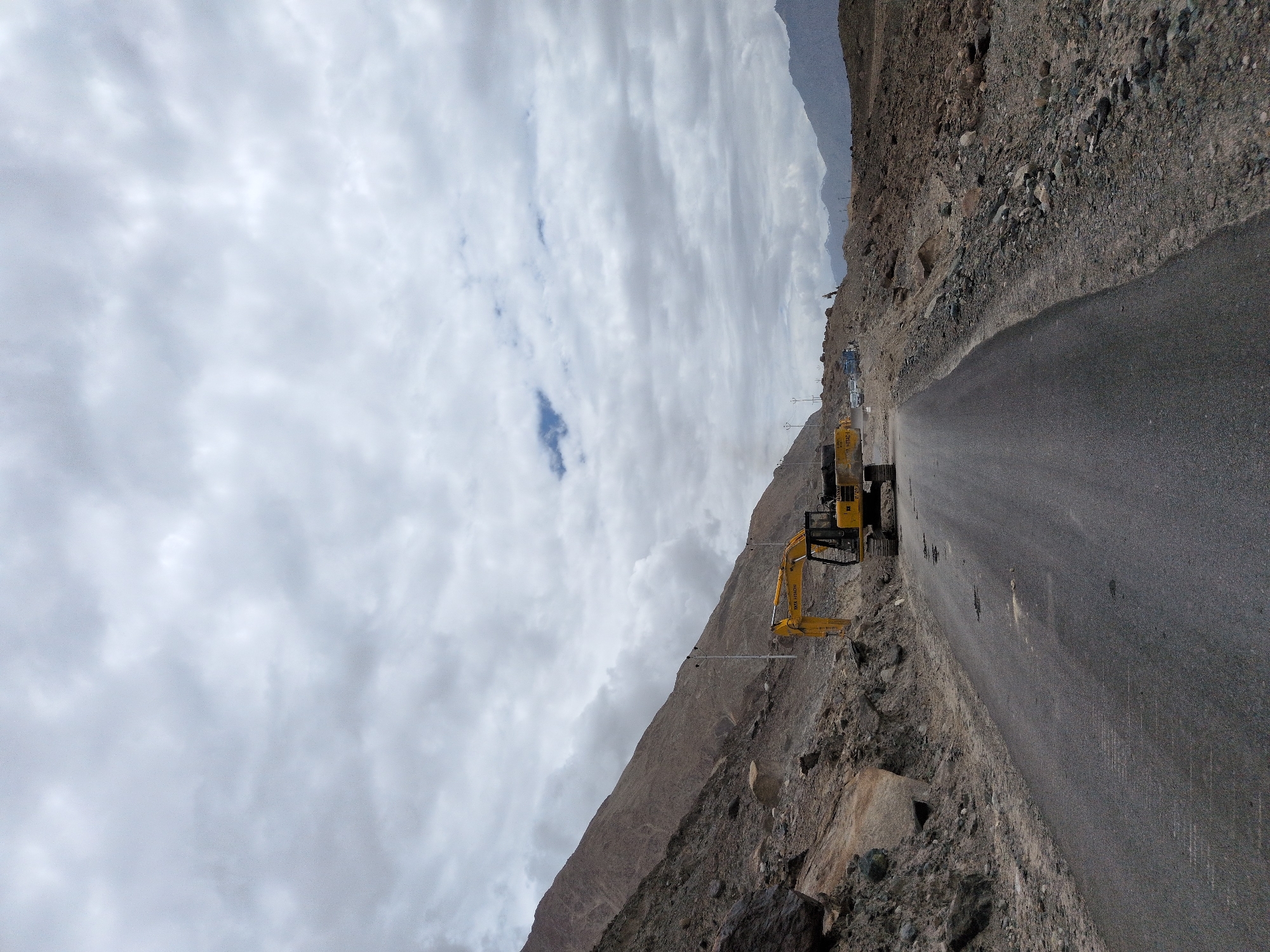 The image depicts a road construction site in Panamik, India. A yellow excavator is positioned on the right side of the road, with its arm extended. The road itself appears to be paved but has some potholes. To the left of the road, a steep, rocky mountainside rises, showing signs of excavation and debris. The landscape is barren and mountainous, with sparse vegetation. The sky is overcast with heavy clouds, suggesting a cloudy or potentially rainy day. In the background, more mountains are visible in the distance, shrouded in haze. There are a few utility poles along the side of the road. A small blue patch of sky is visible amidst the clouds. The scene conveys a sense of ongoing development or repair work in a rugged, remote environment.