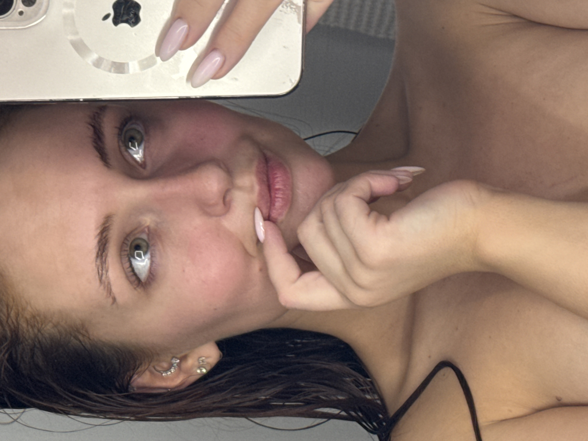 A woman is taking a selfie with an iPhone, covering the top portion of her face with the phone. She is light-skinned with brown hair. The woman has green eyes, manicured nails, and is wearing ear piercings. Her right hand is touching her face. She appears to be topless. The location is unknown and there is no information to suggest that the image was taken in Chaikovsky, Russia.