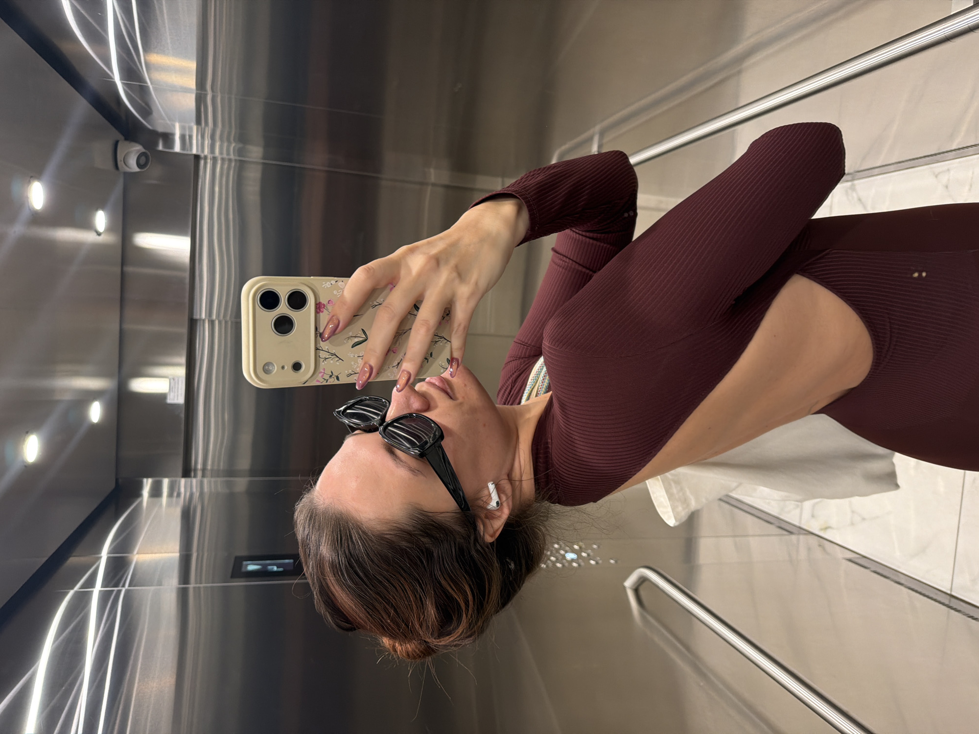 A young woman is depicted taking a selfie inside a modern elevator with highly reflective, polished metal walls. The image is oriented sideways, showing the woman's profile as she holds a smartphone with a floral-patterned case up to capture her reflection. She has dark hair styled in a bun and wears black sunglasses resting on her head, along with a white wireless earbud in her left ear. Her nails are manicured with an ombré pink and purple design. She is dressed in a form-fitting, ribbed, maroon long-sleeved top or bodysuit featuring a cut-out detail on her side.
The elevator interior is sleek, with recessed circular lights illuminating the metal walls, creating bright streaks from reflections. A small white security camera is mounted in the upper left corner of the elevator. A silver handrail is visible on the right side of the frame. The floor appears to be a light-colored, possibly tiled surface. The woman's expression is neutral, focused on the act of taking the photo. There is no discernible text visible in the image.
