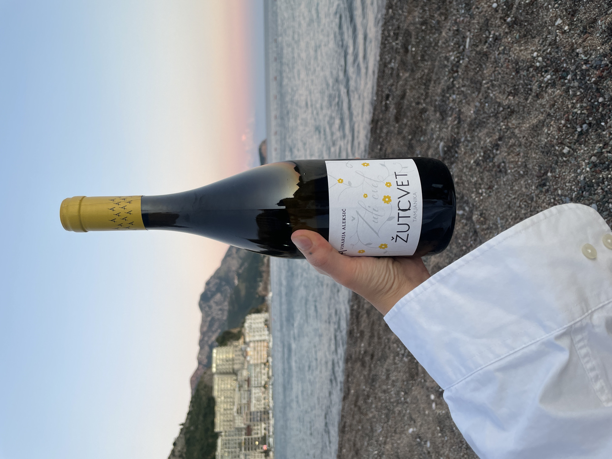A hand in a white shirt holds a bottle of wine on a beach in Rafailovići, Montenegro. The bottle has a yellow cap with an all-over pattern and a white label with text and yellow flowers. The beach is composed of small dark pebbles. In the background, the calm sea meets the shore, and across the water, a city with white buildings is visible, nestled at the base of green, mountainous terrain. The sky is a gradient of pale blue and pink, suggesting either sunrise or sunset.
