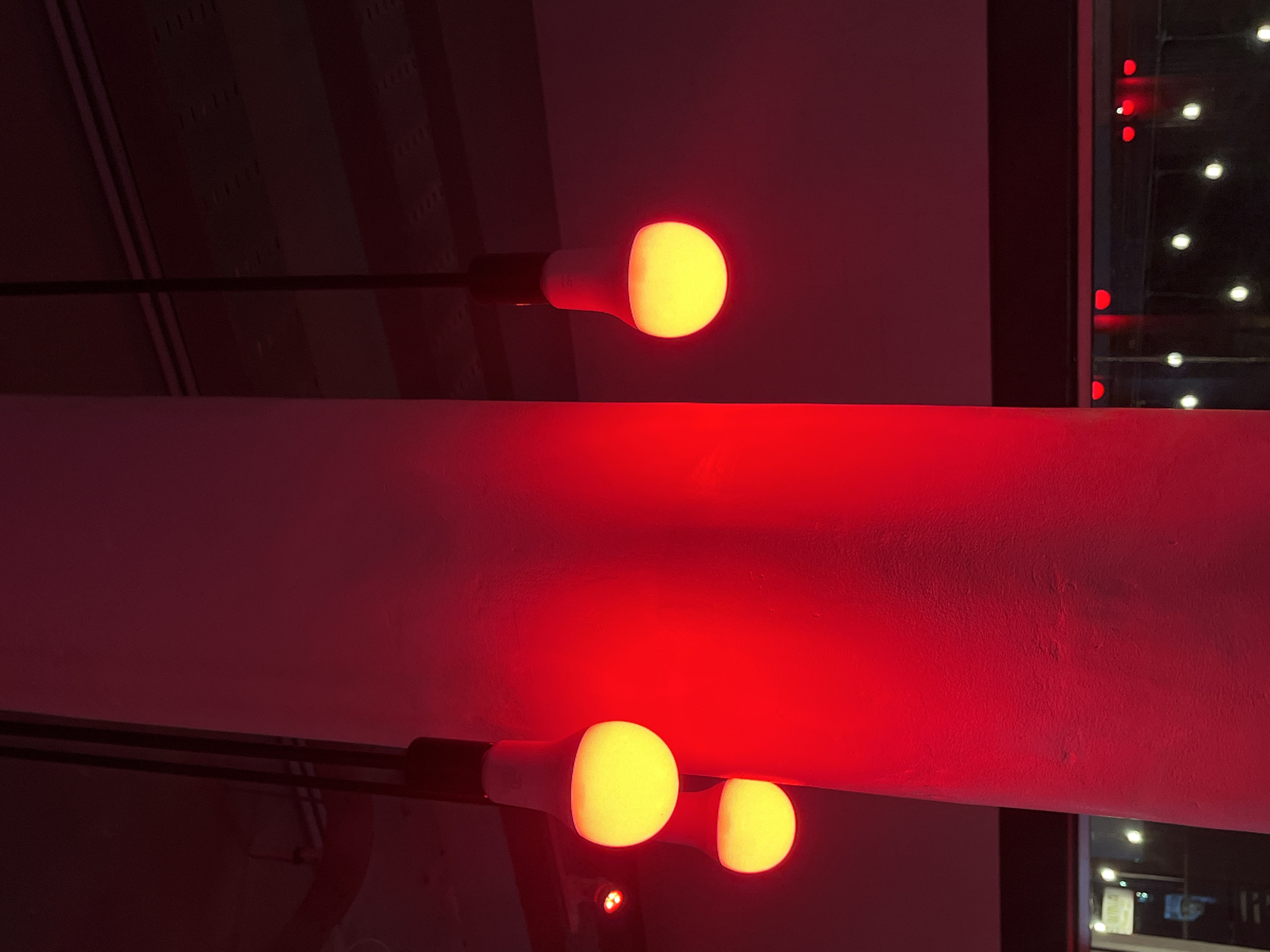 The photograph, taken in North Kuta, Indonesia, features a red-lit interior. The scene is dominated by a bright red light source reflecting off a horizontal surface, likely a beam or architectural element. In the foreground, two light bulbs with yellow-white glows are visible, positioned near the red-illuminated surface. One light bulb is partially obscured. A third light bulb, similar in appearance, is located in the upper portion of the frame and is connected to a black pole. To the right, a window displays a nighttime scene with several lights visible in the distance. The overall lighting conditions cast a strong red hue across the entire setting.