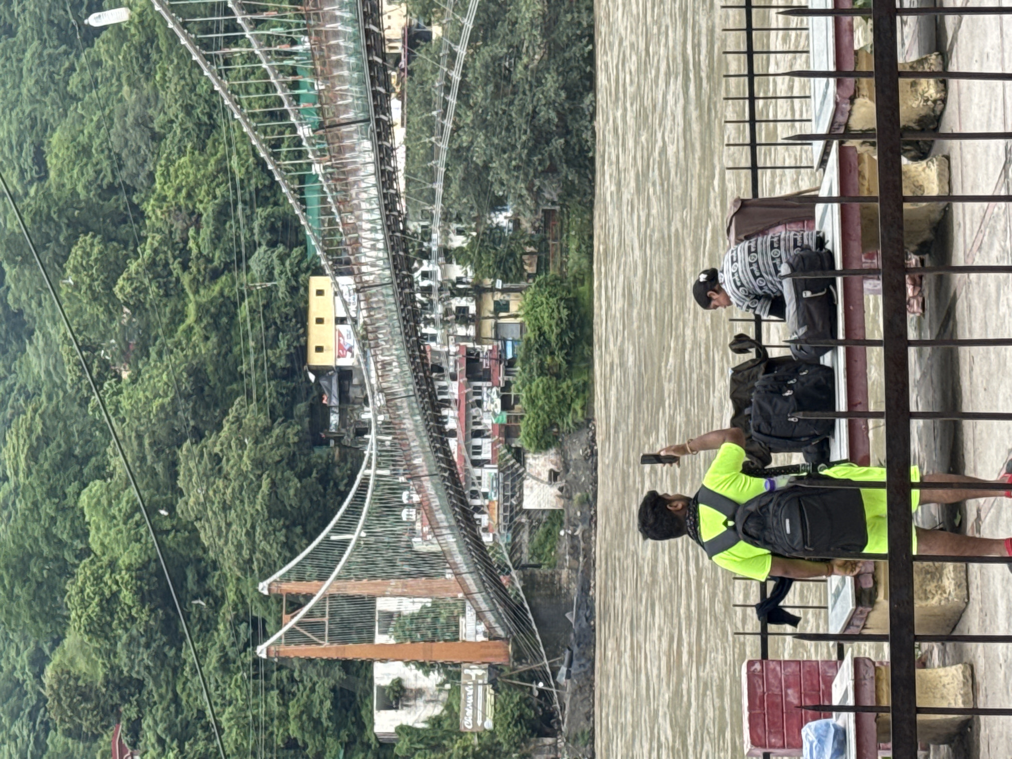 In Rishikesh, India, a man wearing a neon green t-shirt and shorts, with a black backpack, stands facing away from the camera, holding a smartphone up as if to take a picture. Beside him, a woman wearing a patterned sweater sits facing away from the camera, with a backpack and bag placed in front of her. They are positioned on a concrete platform, with a metal fence in the foreground and a wide, murky river behind them. In the background, a suspension bridge stretches across the river, supported by orange towers. The bridge is lined with buildings on its far side, and a lush green hillside covered in trees rises behind the structures. The sky is overcast.