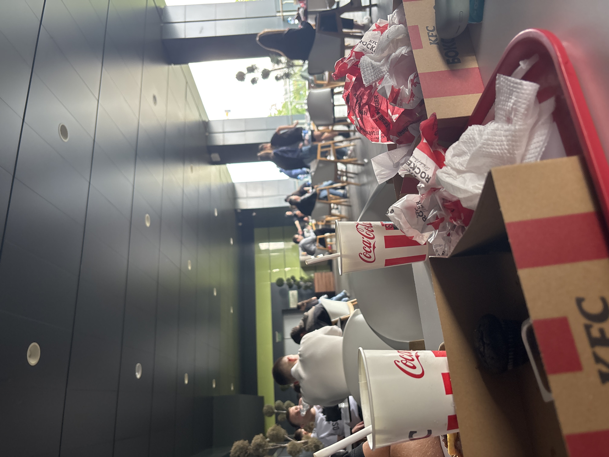 The image shows an interior view of what appears to be a KFC restaurant. A red plastic tray is in the foreground, cluttered with KFC boxes and crumpled wrappers, alongside two Coca-Cola cups with straws. The packaging suggests the meal includes burgers and other KFC items. In the background, patrons are seated at tables, possibly dining or waiting for their orders. The setting includes black walls and rows of chairs and tables. Light enters through windows and doorway. The location is in the city of Plovdiv, Bulgaria.