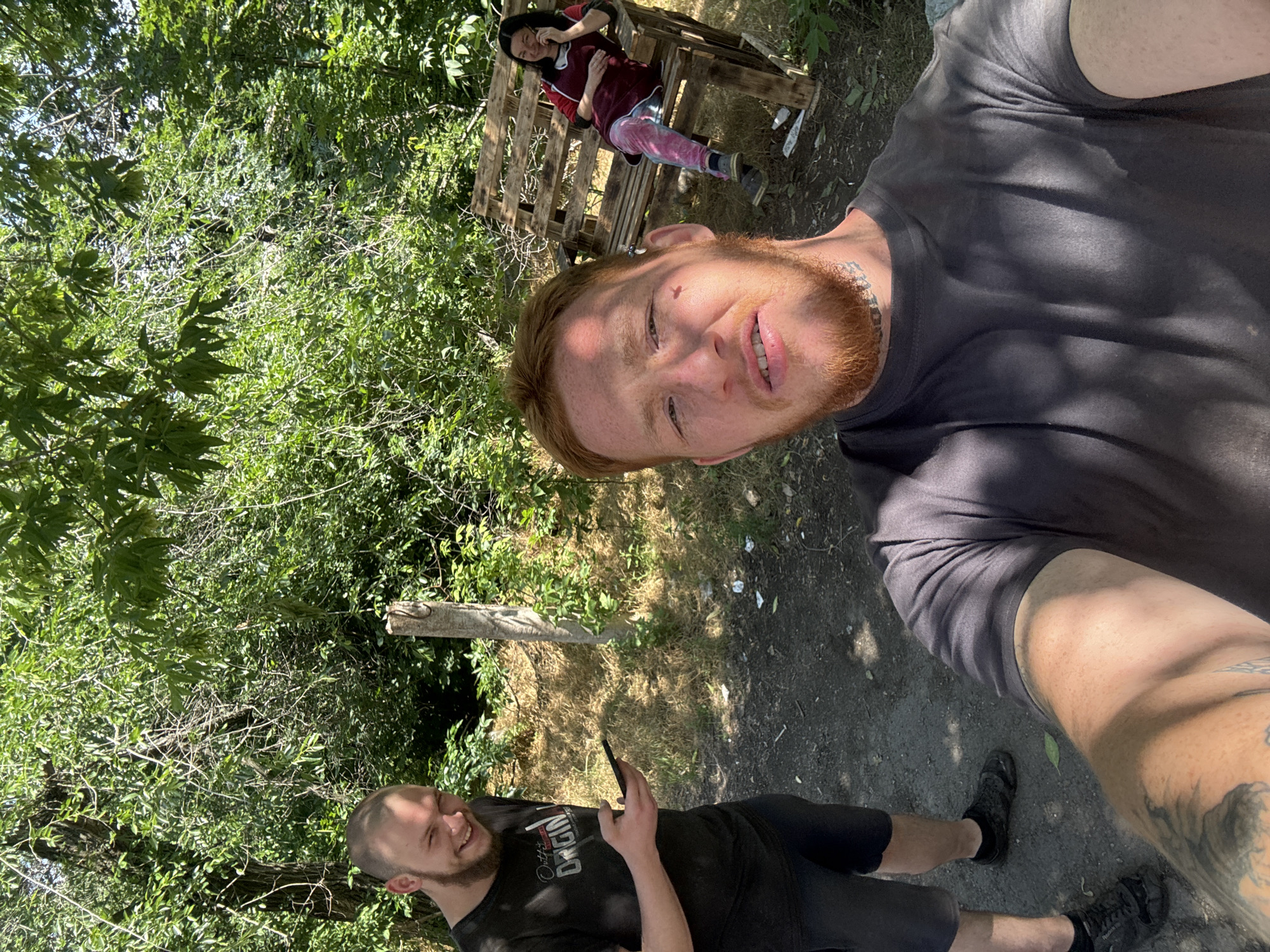 In a wooded area, three individuals are present. In the foreground, a person with red hair and a beard is taking a selfie, their face partially obscured by the camera. To the left, a man with a shaved head and a beard smiles while holding a phone. In the background, a woman sits on a wooden bench, talking on her phone. The environment is characterized by dense green foliage, dry grass, and dirt ground. The overall scene appears casual and informal.