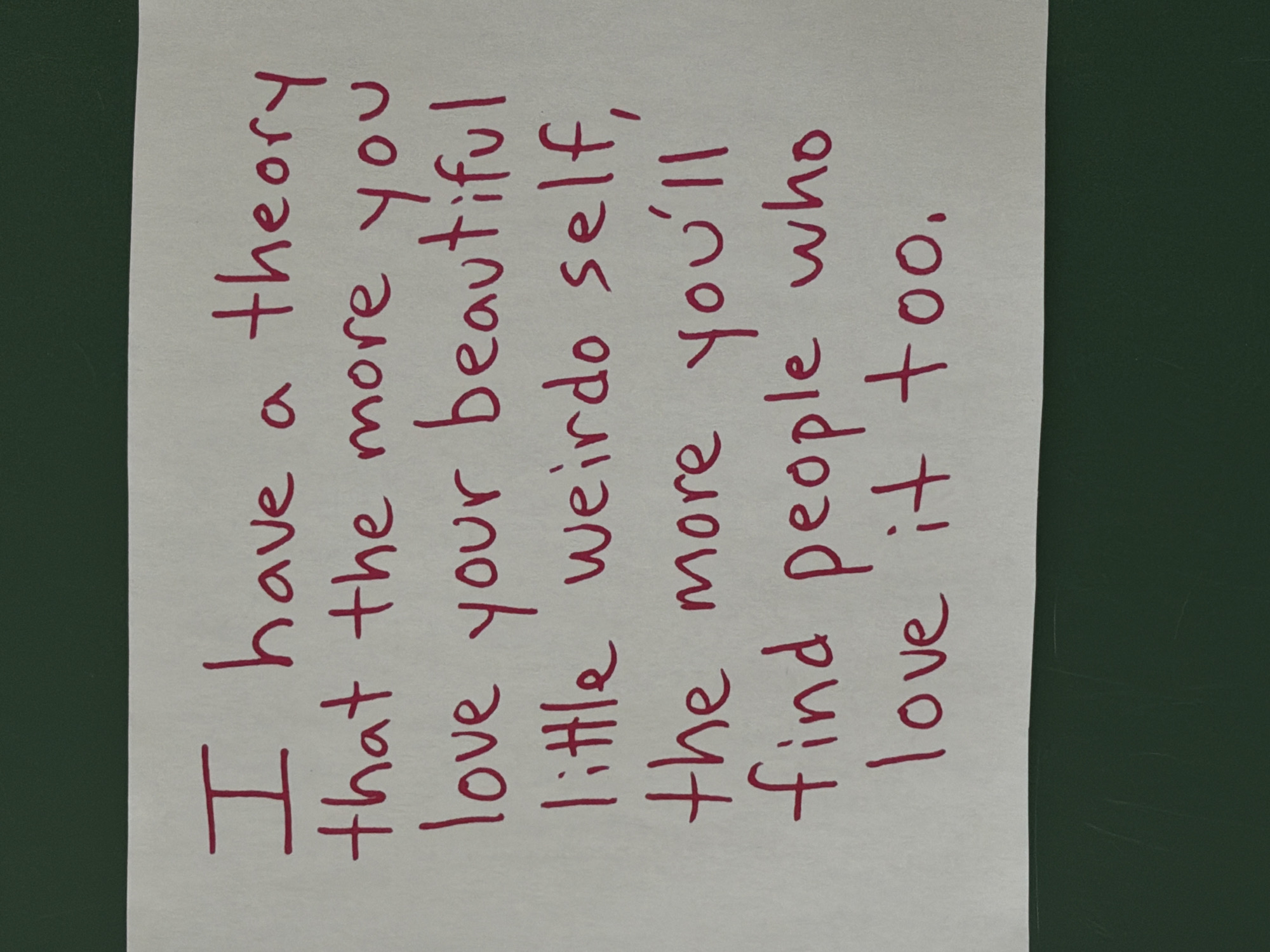 A photograph shows a handwritten note with maroon ink on white paper. The note reads, "I have a theory that the more you love your beautiful little weirdo self, the more you'll find people who love it too." The paper rests on a dark green surface. The location and city are unknown.
