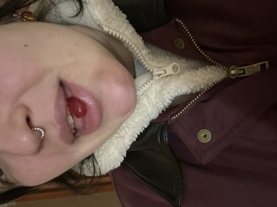 A close-up image depicts a person from the chin to just above the eyes, positioned slightly off-center. The individual has a light complexion and dark hair visible on the left side of the frame. A septum piercing, a silver-colored hoop with small clear gems, is present. An amber-colored earring hangs from the right earlobe. The person's mouth is slightly open, revealing a small, red, translucent, irregularly shaped object, possibly a candy or fruit, held by the tongue against the inside of the lower lip. The lips are full and appear glossy. The individual wears a maroon or burgundy jacket featuring a dark, possibly faux leather, collar and a visible white, fuzzy sherpa lining around the neckline. A metal zipper pull is evident on the partially unzipped main zipper of the jacket, and brass-colored snap buttons are visible on the jacket's front flap. A light gray garment is partially visible beneath the sherpa lining. The background is dark and out of focus, suggesting an indoor environment. The location in the city of Chaikovsky, Russia, cannot be determined from the visual information presented.