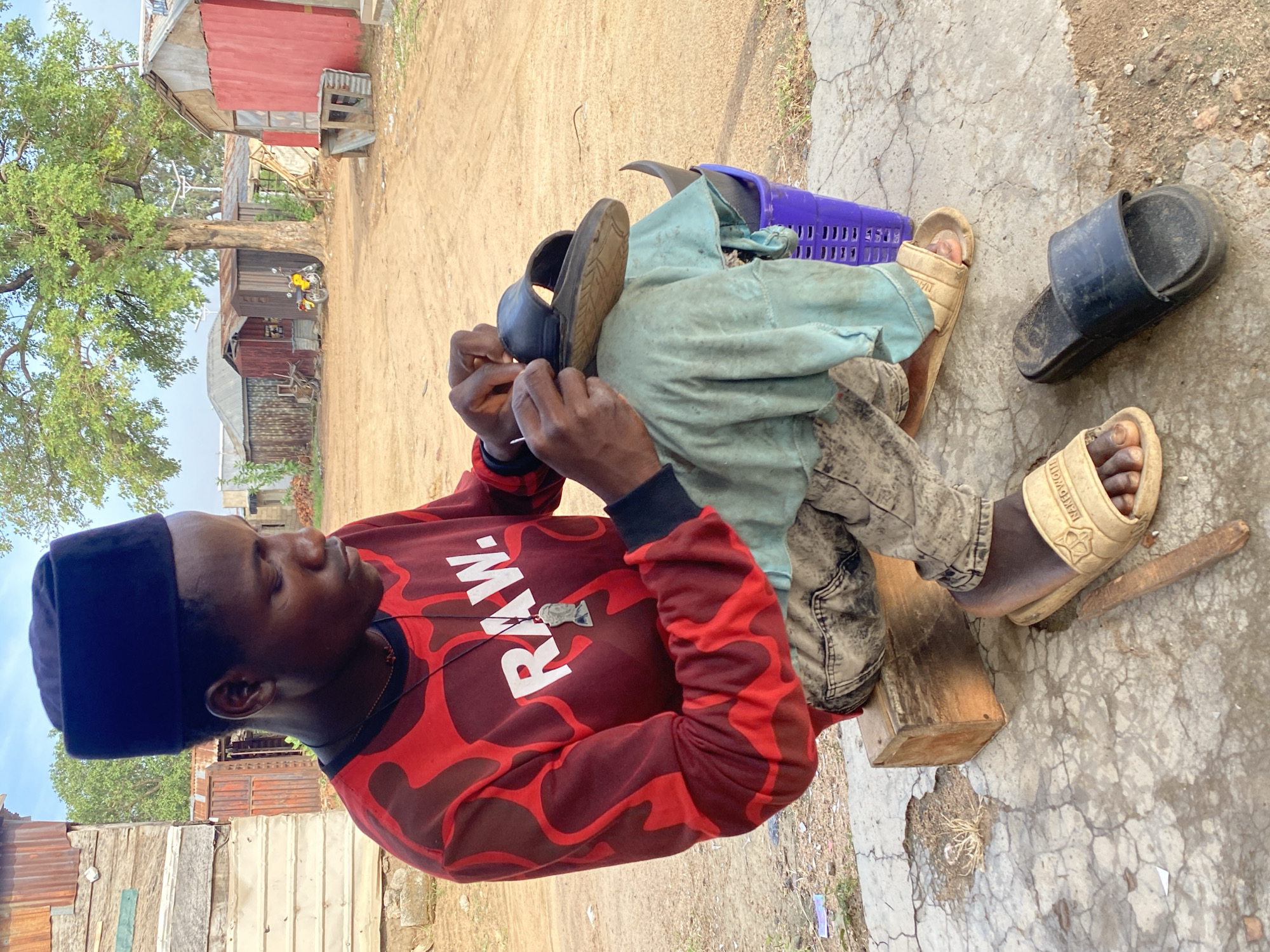 The image depicts a young man intently repairing a sandal outdoors in Zango-ndaji, Nigeria, on a bright, clear day. The man, with dark skin, wears a dark blue flat-topped hat and a long-sleeved maroon and red patterned shirt featuring the white text "RAW." on the front. A silver-colored pendant hangs from his neck. He is seated on a wooden block, using a small tool to mend a dark-colored sandal, demonstrating focus on his task.
Adjacent to him are the lower legs of another person, clad in light blue trousers and light beige slide sandals, one foot positioned near the shoe being repaired. The beige sandals have partially visible text that reads "NAKEDWOL...". Nearby, a purple plastic basket and an additional dark slide sandal rest on the ground, which is a mix of dirt and cracked concrete.
The background reveals a rustic, semi-urban setting with several simple buildings constructed from corrugated metal sheets and wood, including one with a red-painted wall. A large tree with lush green foliage stands prominently, and a yellow motorcycle is parked near one of the structures.