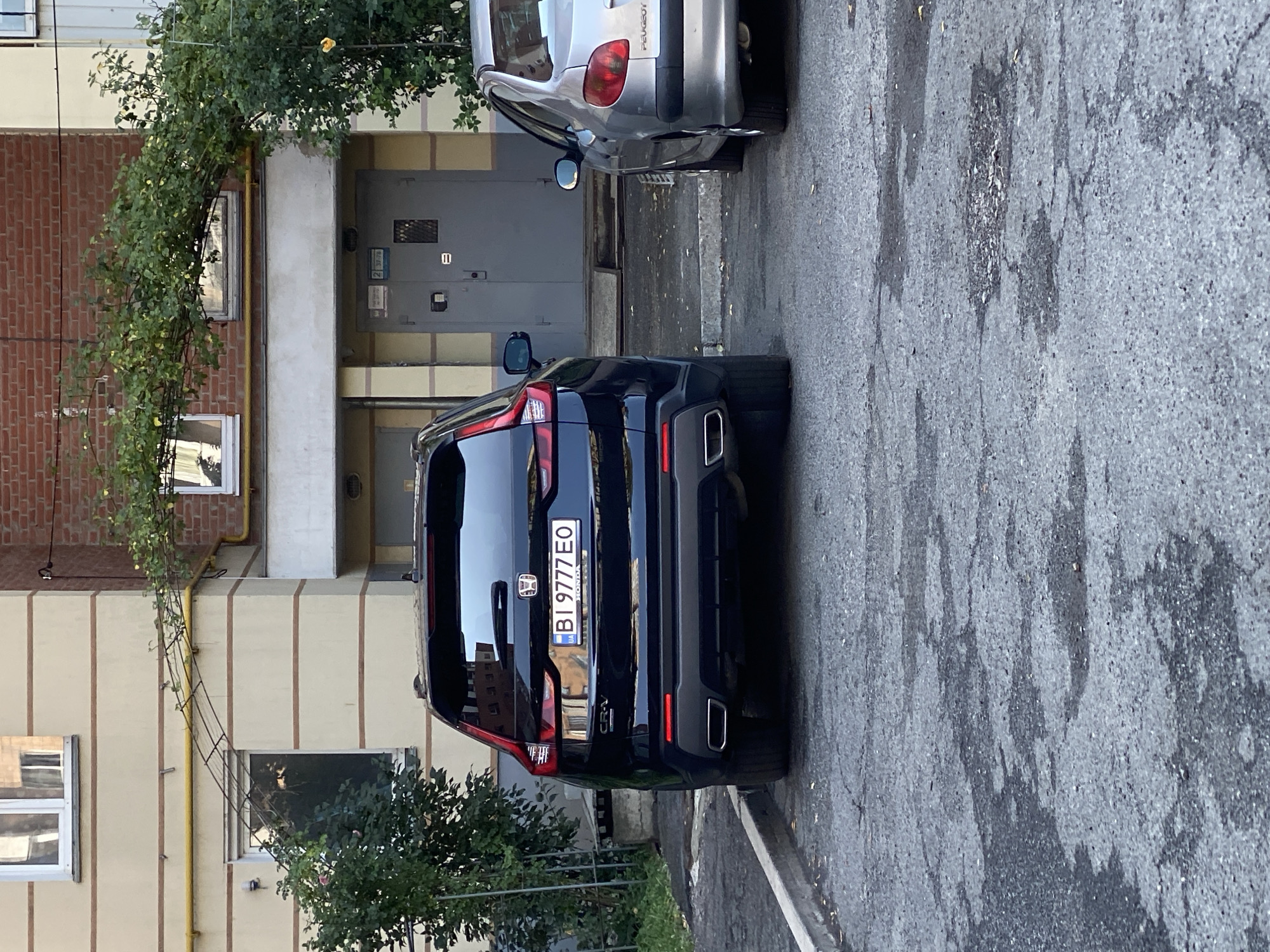 A black Honda CR-V is parked in front of a building with brick and beige siding. The license plate on the car reads "BI 9777 EO". A silver Peugeot is parked to the right of the Honda. The ground is asphalt with some cracks and water stains. There is greenery on the building and in front of it. The location is Poltava, Ukraine.