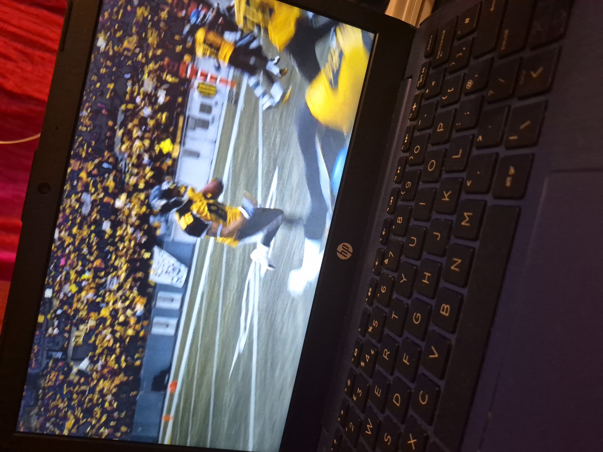 The image shows a close-up view of a laptop screen displaying a football game, likely American football, with players in yellow and black uniforms on a green field. The laptop itself is open and positioned on a dark surface, with a portion of the keyboard visible in the foreground. The keyboard is a standard QWERTY layout, with the HP logo visible on the laptop's bezel. The background is dark, suggesting an indoor setting, with hints of red fabric at the top. There are no discernible landmarks or elements that would suggest a specific location within Sandwell, United Kingdom. The image is focused on the laptop and the game being played.