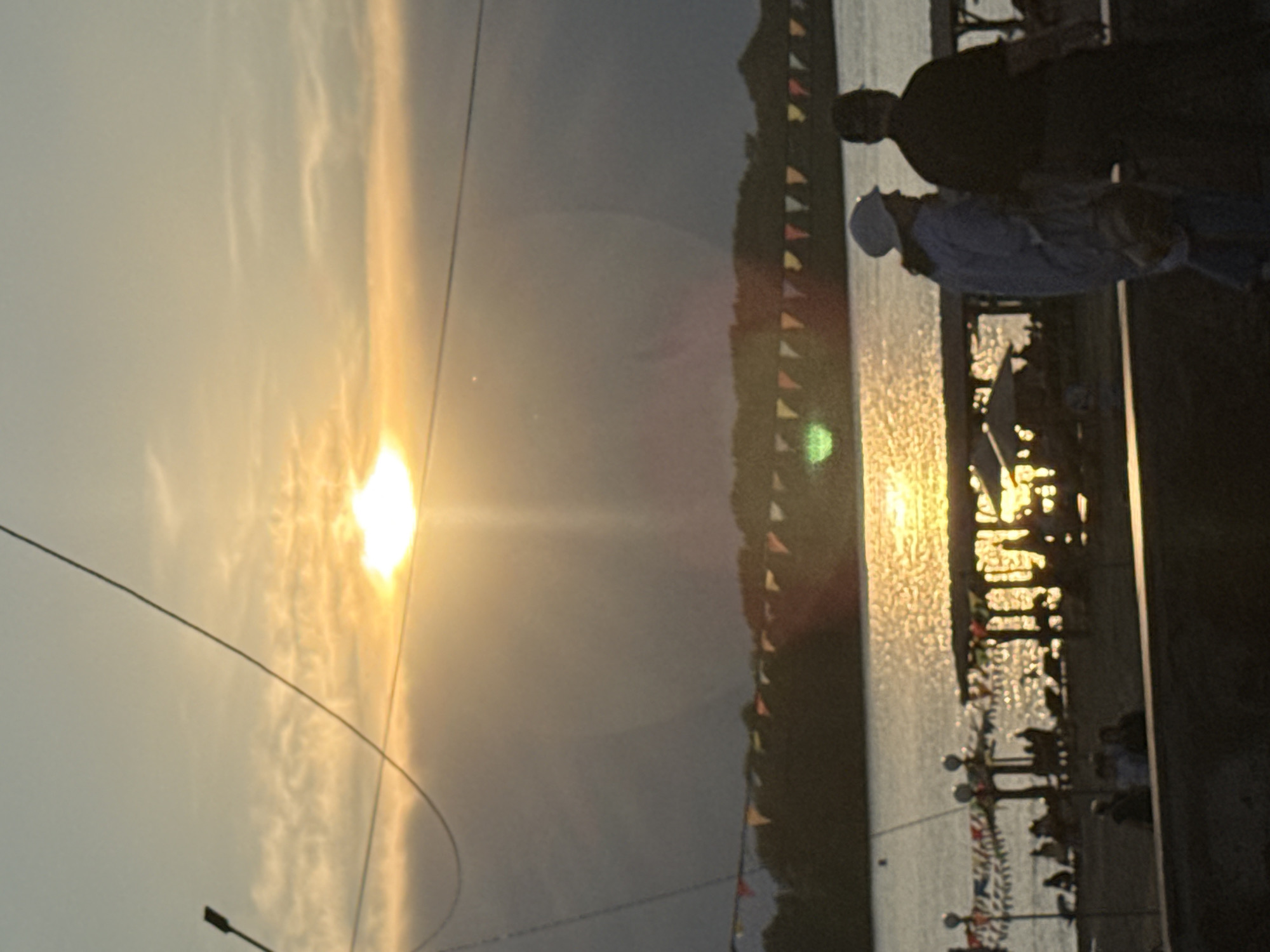 The image depicts a sunset over the Irtysh River in Pavlodar, Kazakhstan. Silhouetted figures can be seen on a beach area with umbrellas and decorative flags strung between poles. The sun is partially obscured by clouds, casting a warm glow on the water. In the foreground, two individuals are walking away from the camera, their features indistinct due to the low light. The overall scene suggests a leisurely evening at a waterfront location.