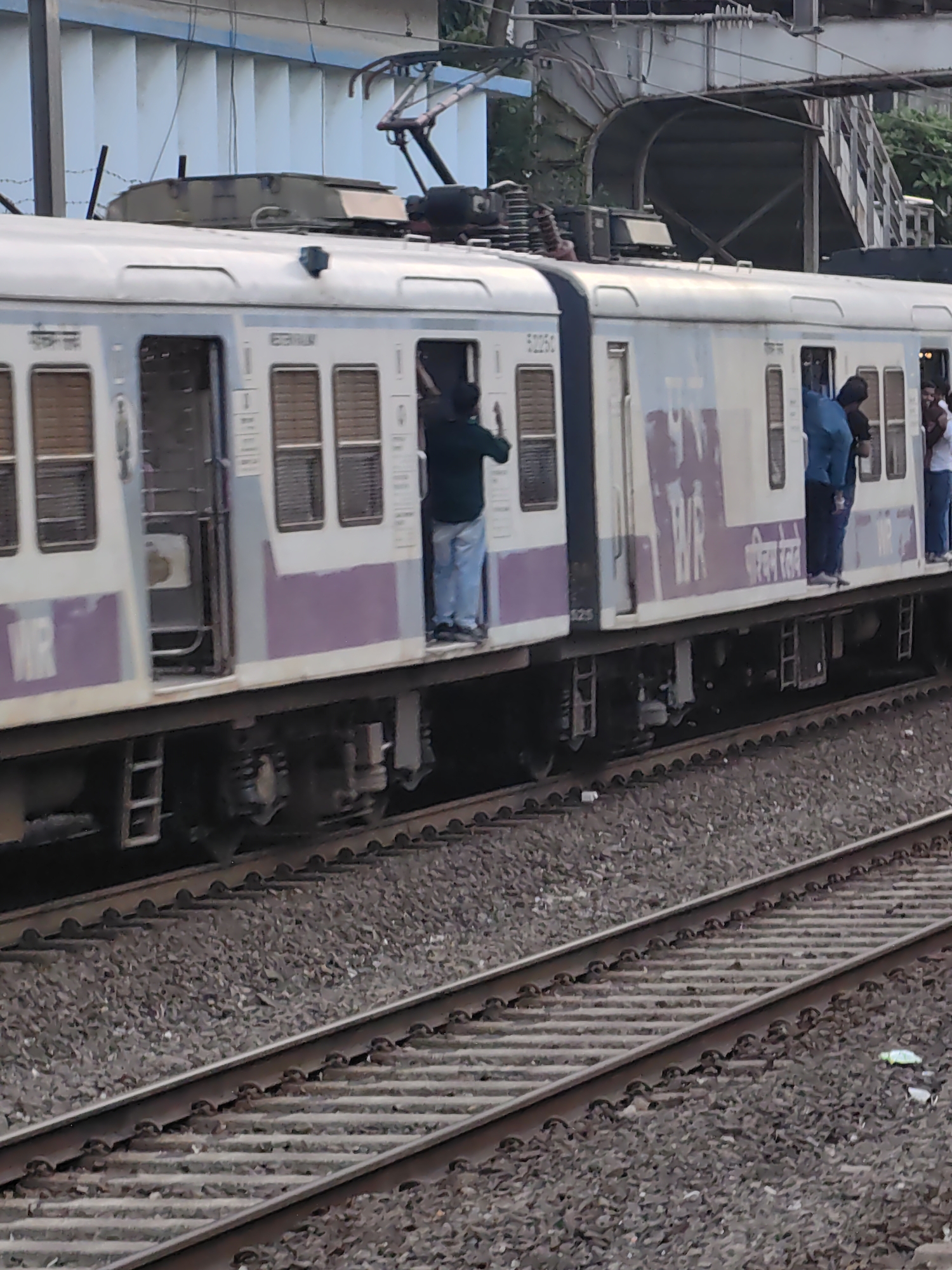 The image shows a suburban train in Mumbai, India, at a station. The train is an electric multiple unit (EMU) with several coaches. Passengers are seen standing on the footboards of the train, with some leaning out of the open doors. The train is on a railway track, with ballast and sleepers visible. In the background, there is a platform and overhead electrical equipment for the train. The sky is overcast. The image appears to be taken from another train or an elevated position. The presence of people hanging on the outside of a moving train suggests a common practice in overcrowded Mumbai local trains, indicating a densely populated urban environment. The train's livery, the presence of overhead wires, and the style of the carriages are characteristic of Mumbai's local train network, which is managed by Western Railway (WR) and Central Railway (CR). The scene is typical of rush hour on the Mumbai suburban railway system.
