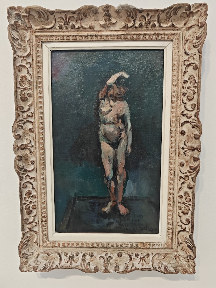 The image displays a vertically oriented oil painting, identified as "La Danseuse" (The Dancer) or "Nu debout" (Standing Nude) by Henri Matisse (c. 1907), housed within the Musée national d'Art Moderne at the Centre Pompidou, Paris, France.
The painting depicts a full-length, standing nude female figure, rendered with a simplified form in tones of pale pink, orange, and reddish-brown, accentuated by dark outlines and greenish-blue shadows. Her left arm is raised with the hand positioned near or on her head, while her right arm hangs alongside her body. The figure stands upon a dark, angular surface, against a mottled, dark teal or greenish-blue background. The artist's signature, "HENRI MATISSE," is legible in dark script in the lower right corner of the canvas.
The artwork is enclosed by an ornate, off-white, distressed wooden frame featuring intricate carved scrollwork and acanthus leaf motifs. A plain white inner liner separates the frame from the painted canvas. The framed painting is centrally mounted on a smooth, uniformly lit, plain white wall, consistent with a museum or gallery display environment.