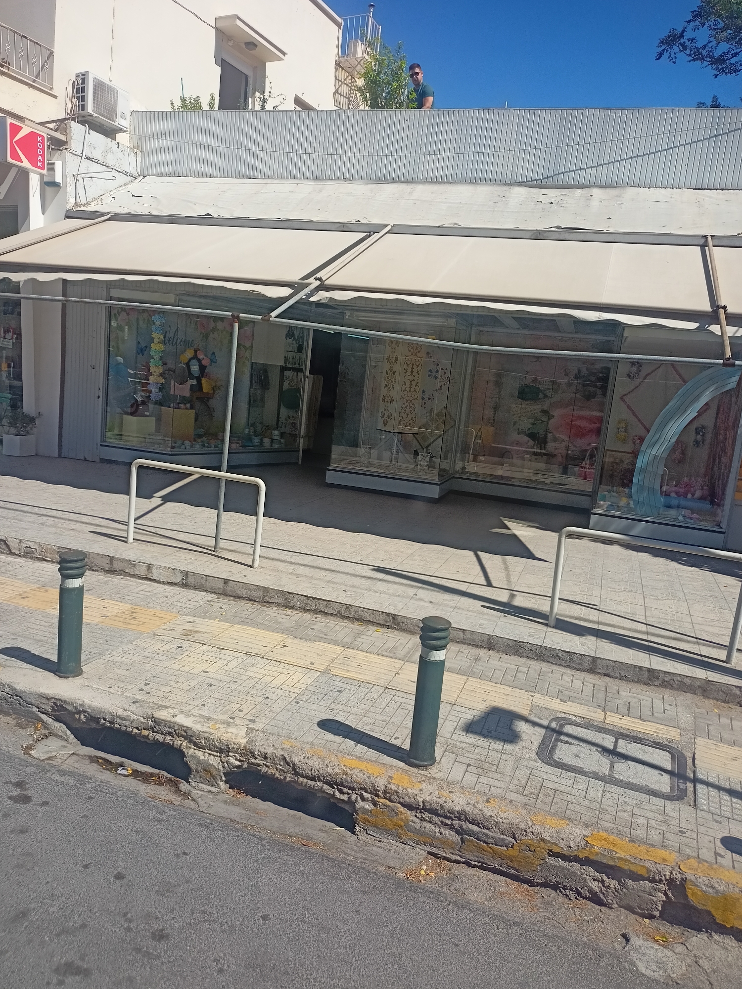 The image captures a street scene in an unknown city. In the foreground, the asphalt road meets a raised sidewalk constructed with light-colored pavers. A green bollard stands on the sidewalk. A damaged curb, painted yellow on top, separates the road from the sidewalk. The sidewalk is further delineated by a white metal railing. Behind the railing, a shop with large glass windows is visible, displaying various items. The shop has a cream-colored awning extending over the storefront. Above the awning, a portion of a building is visible, with an air conditioning unit mounted on its exterior. On the rooftop of the building, a person wearing a dark green shirt is standing and looking towards the camera. The sky is clear and blue.