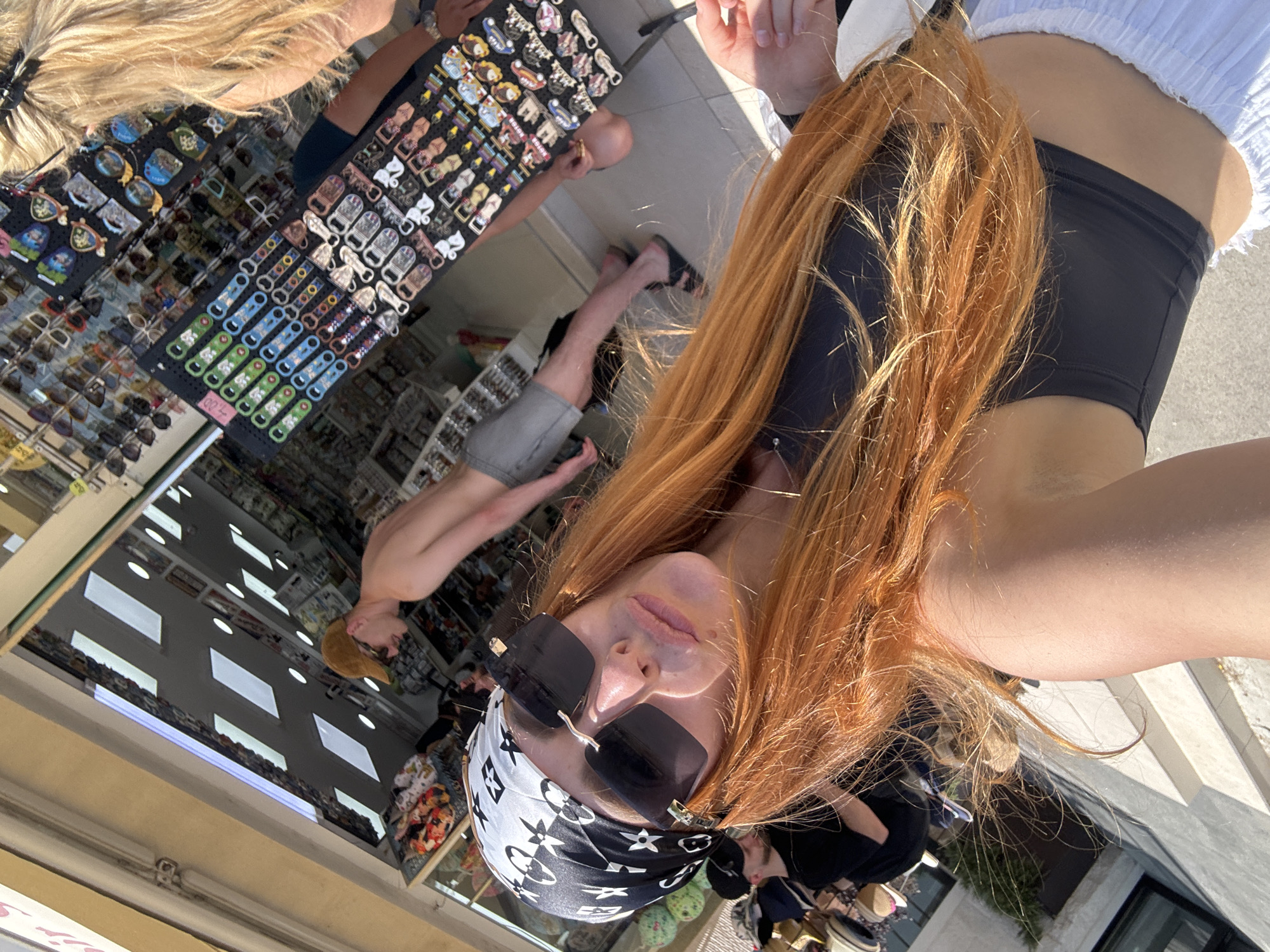 In Budva, Montenegro, a woman with long, red hair and a bandana tied around her head is taking a selfie in front of a shop. She is wearing sunglasses and a dark top. Behind her, a shirtless man wearing a brown cap and grey shorts is browsing items displayed on a rack. The shop appears to be selling souvenirs and accessories, with racks of sunglasses and keychains visible. The scene is outdoors, with sunlight casting shadows on the ground.