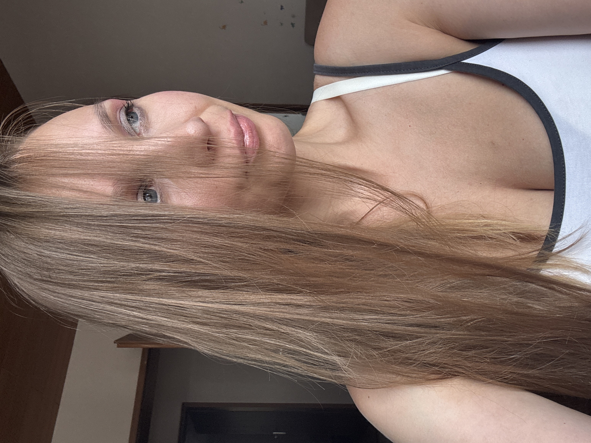 A woman with long, light brown hair is photographed from the chest up, looking to her right. Her hair partially obscures her face, with strands falling across her forehead and over her left eye. She is wearing a white tank top with dark grey trim. Her skin is fair and appears to be lightly made up. The background is dimly lit and indistinct, with a suggestion of interior walls and possibly a doorframe. The image does not provide any visual information that can be used to determine the location within the city of Überherrn, Germany.