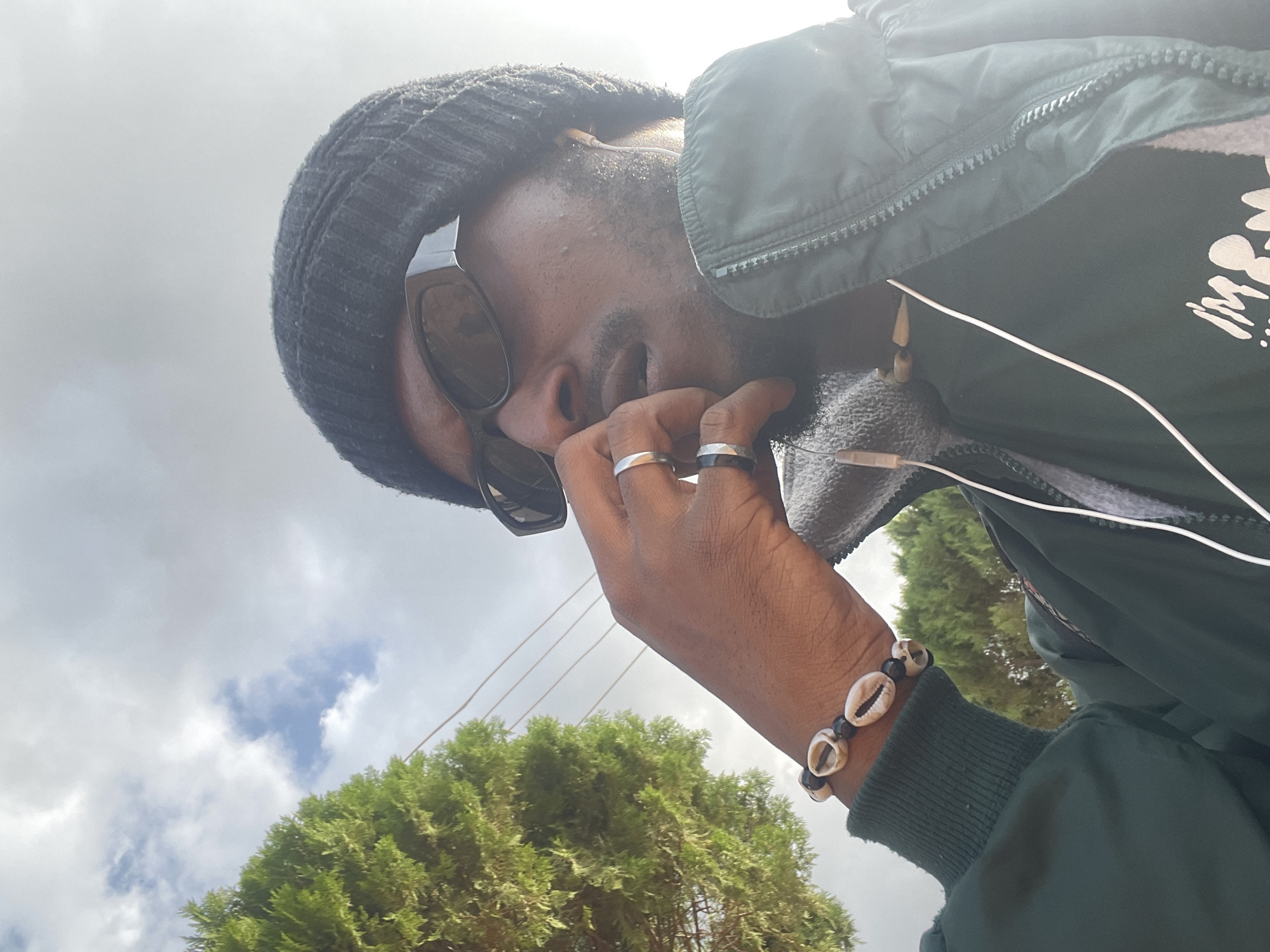 A medium shot captures a person from the chest up, outdoors, with a tree on the left and a bright, cloudy sky above. The person, viewed from a low angle, wears a black beanie, dark sunglasses, and a dark green jacket over a gray hooded sweatshirt. They have on their right wrist a bracelet made of white shells and black beads, and a second bracelet of similar design is visible further down the arm. Two rings are worn on the fingers of the right hand, which is positioned near their mouth. White earbuds are worn, with wires visible. The background suggests an urban setting in Jos, Nigeria, possibly near a residential area or park, given the presence of the tree and power lines. The lighting indicates it is daytime, with the sun creating a bright, hazy effect.