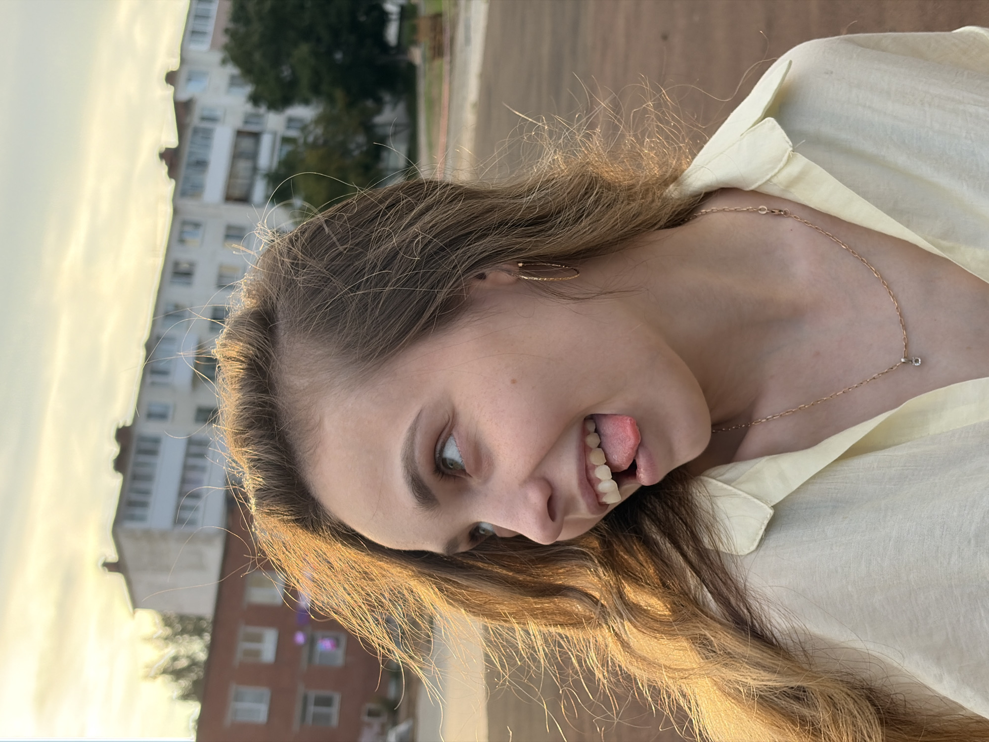 A young woman is captured in a profile shot outdoors, with buildings and trees in the background, suggesting an urban or suburban setting, possibly in Noviy, Russia, given the architectural style of the buildings which appears consistent with Soviet-era apartment blocks. The woman, with long, wavy blonde hair, is looking to her left, with her tongue playfully sticking out. She is wearing a light yellow collared shirt and a delicate gold necklace with a small pendant. The lighting suggests it is late afternoon or early evening, with warm, golden sunlight illuminating her hair and face. The ground in the foreground is a reddish-brown, possibly a sports field or paved area.