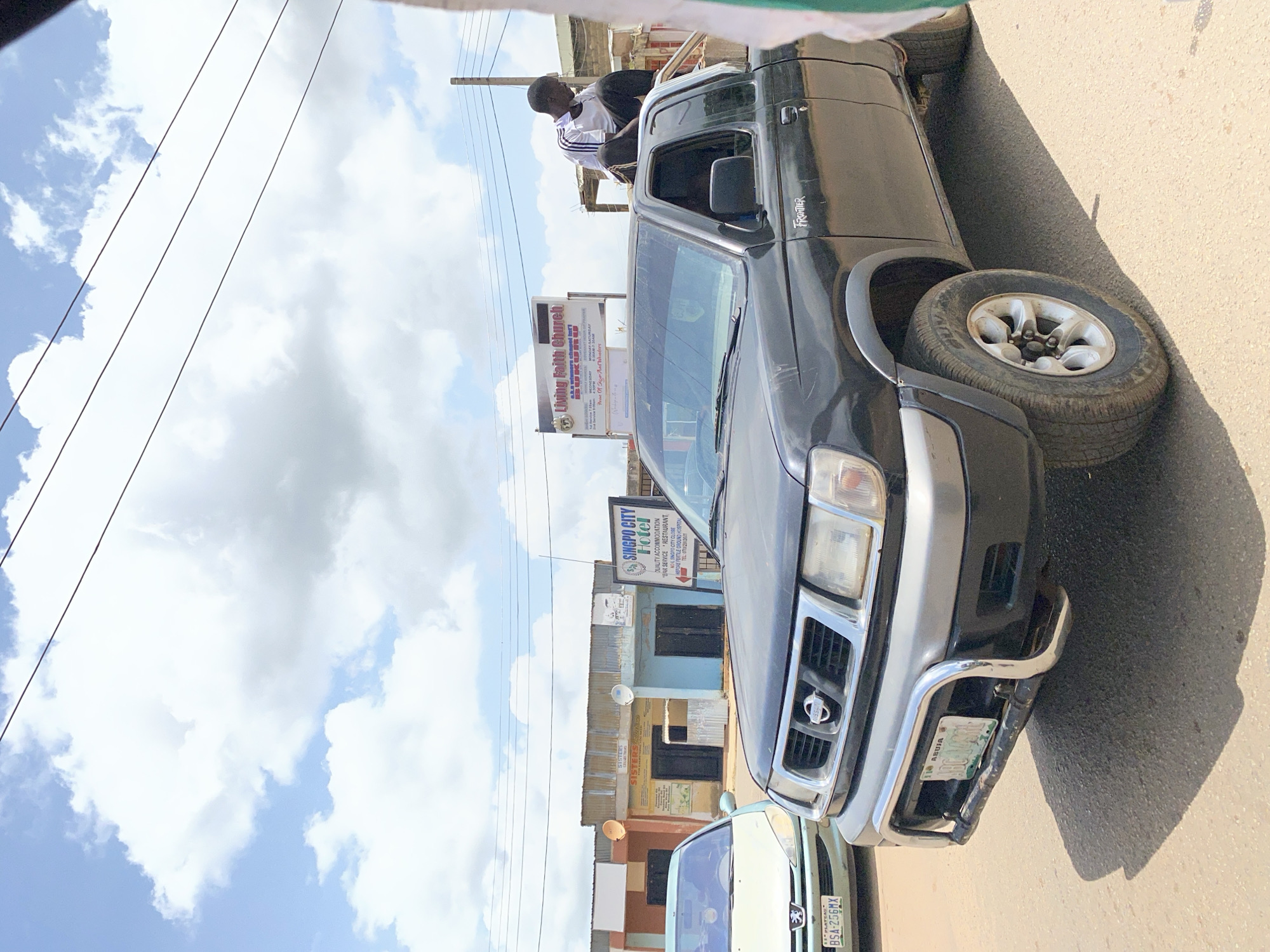 In the city of Gura Top, Nigeria, a black Nissan Frontier pickup truck is parked on a dusty street. A young man, wearing a white and black striped t-shirt and black pants, is sitting on the roof of the truck, looking to the right. In the background, a light blue building with a sign that reads "SINGPO CITY HOTEL" is visible, along with other shops. A light-colored sedan is parked to the left of the pickup truck. The sky is filled with white, fluffy clouds. The scene appears to be in an open area with a few structures and a road.