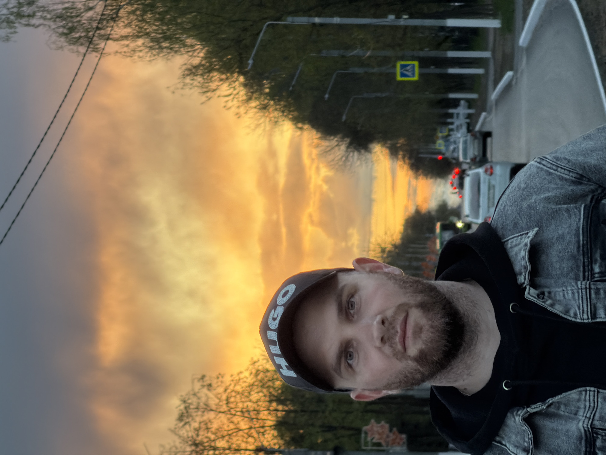 A man is standing on a sidewalk, facing the camera with the city street behind him. He is wearing a black hooded sweatshirt and a gray denim jacket, and a dark baseball cap with "HUGO" emblazoned on it. The street, running behind the man, shows several cars and crosswalk signs. The sky is an orange and yellow color, indicating it is either sunrise or sunset. The sidewalks are concrete. There are trees on both sides of the street. Based on available information, this is the city of Chaikovsky, Russia.