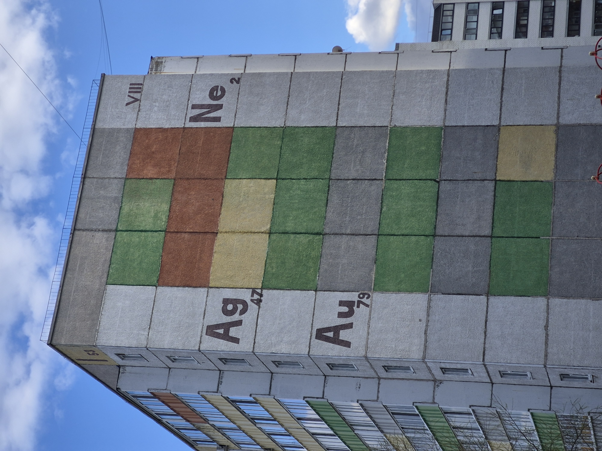 The side of a high-rise building in Tobolsk, Russia is adorned with a depiction of elements from the periodic table. "VIII" and "Ne²" are on the top portion of the building's facade. Below these is a grid of colored squares in rust, yellow, green, and grey. At the bottom of the painted grid are the chemical symbols and atomic numbers for silver and gold, labeled "Ag⁴⁷" and "Au⁷⁹". Windows are visible on the lower floors of the building, and a second building is visible in the upper right corner. A blue sky with clouds is in the background.